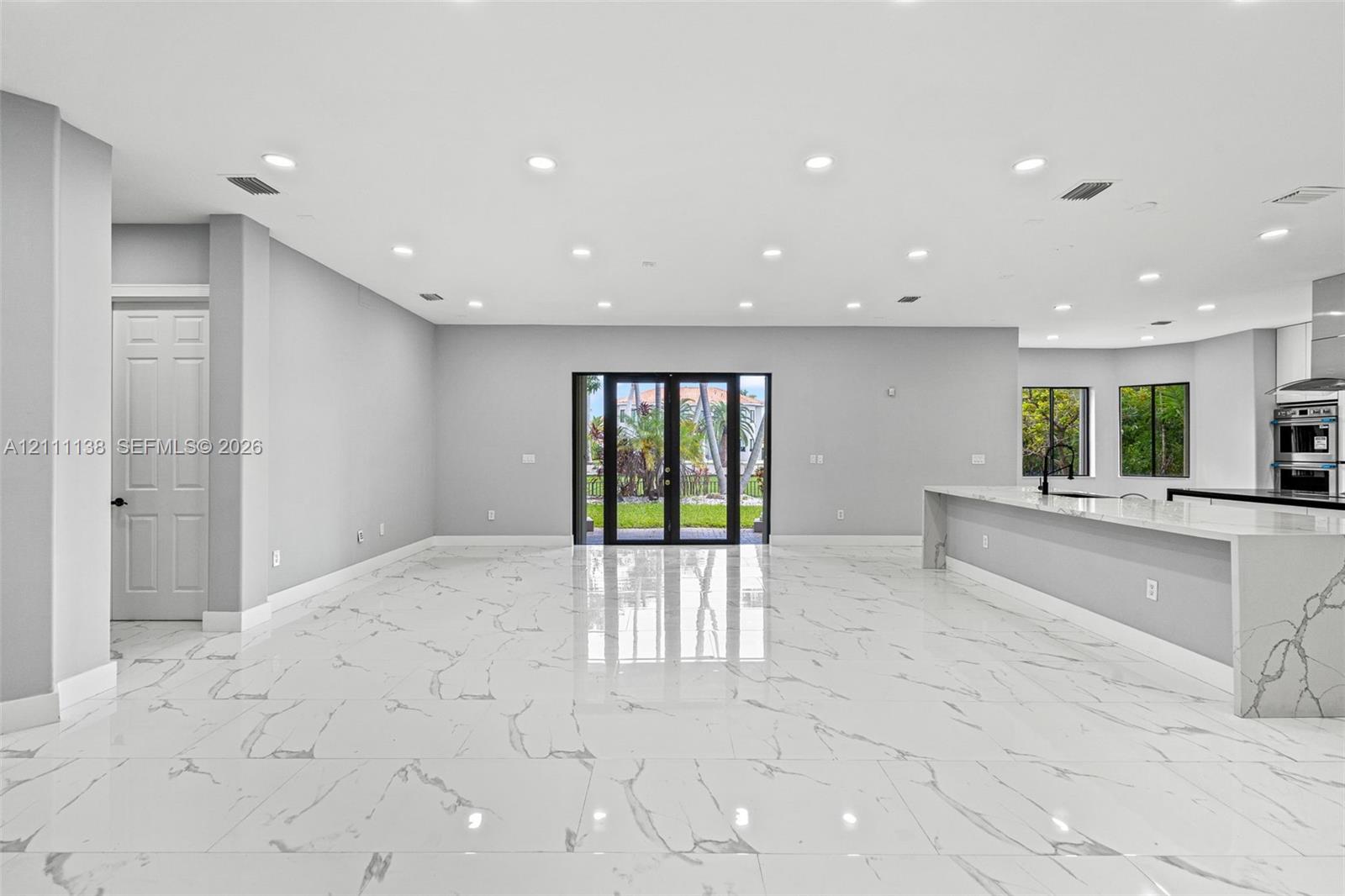 Open-concept interior featuring large-format polished stone flooring, recessed ceiling lighting, a waterfall-edge kitchen island, and double glass-paned doors