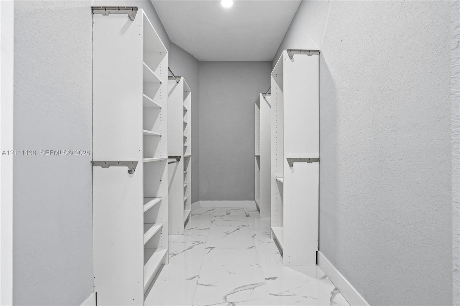 Walk-in closet featuring white built-in shelving, multiple hanging rods, and large-format tile flooring with a neutral vein pattern