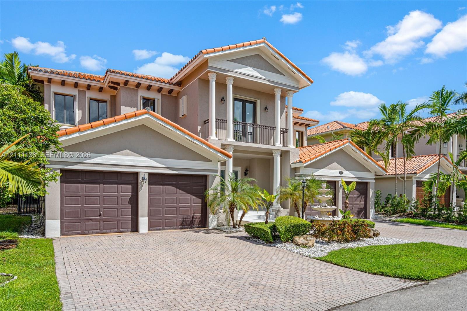 Two-story stucco exterior featuring a tiled roof, multi-bay garage, covered entryway with classical columns, second-level balcony, and decorative stone fountain