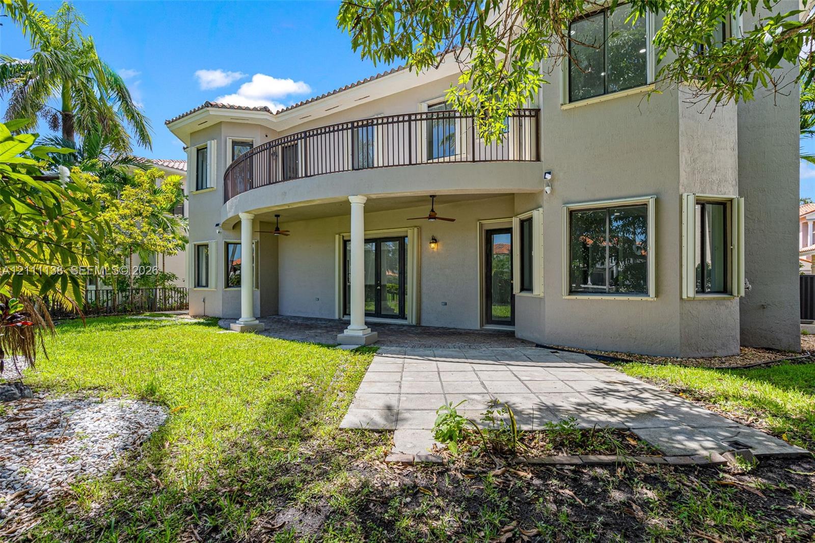 Stucco exterior featuring a covered patio, paved hardscaping, a second-story curved balcony with metal railing, and structural columns
