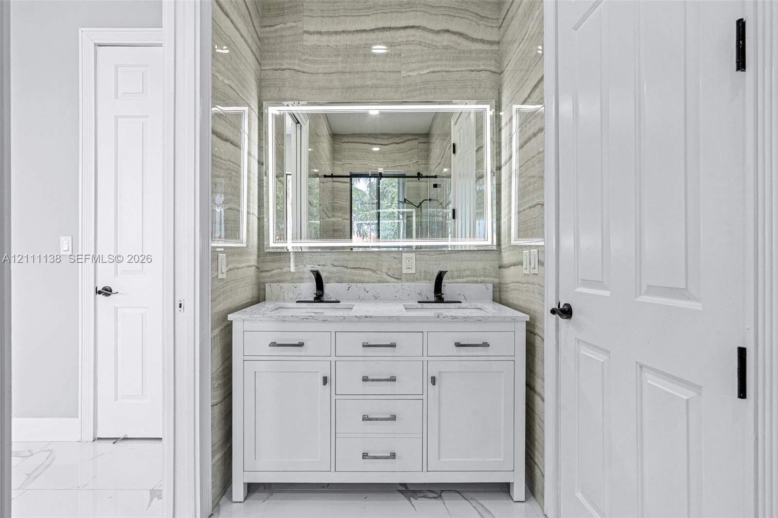 Double vanity featuring a white cabinetry base, stone-look countertop, integrated dual sinks, and matte black widespread faucets, accented by full-height linear pattern tile walls and a modern LED-backlit mirror
