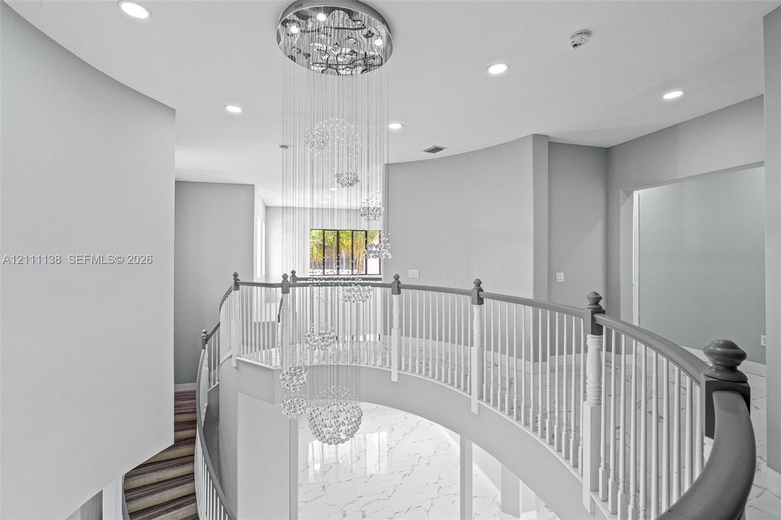 Curved balcony featuring a multi-tiered crystal chandelier, white stone-finish flooring with grey veining, and a white balustrade with a contrasting handrail