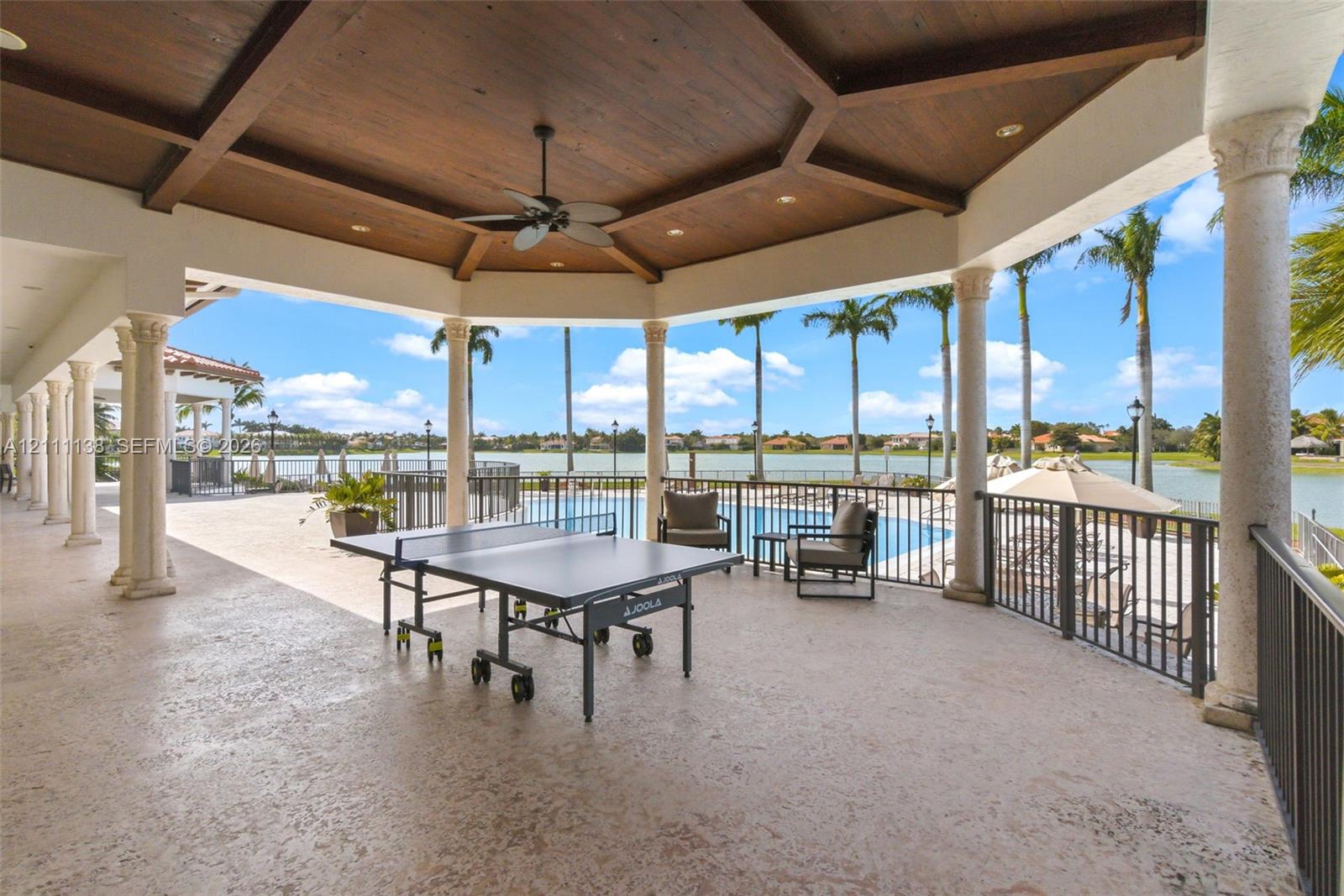 Covered outdoor patio featuring a wood-paneled coffered ceiling, stone-finished flooring, decorative columns, and a metal railing overlooking a pool and waterfront