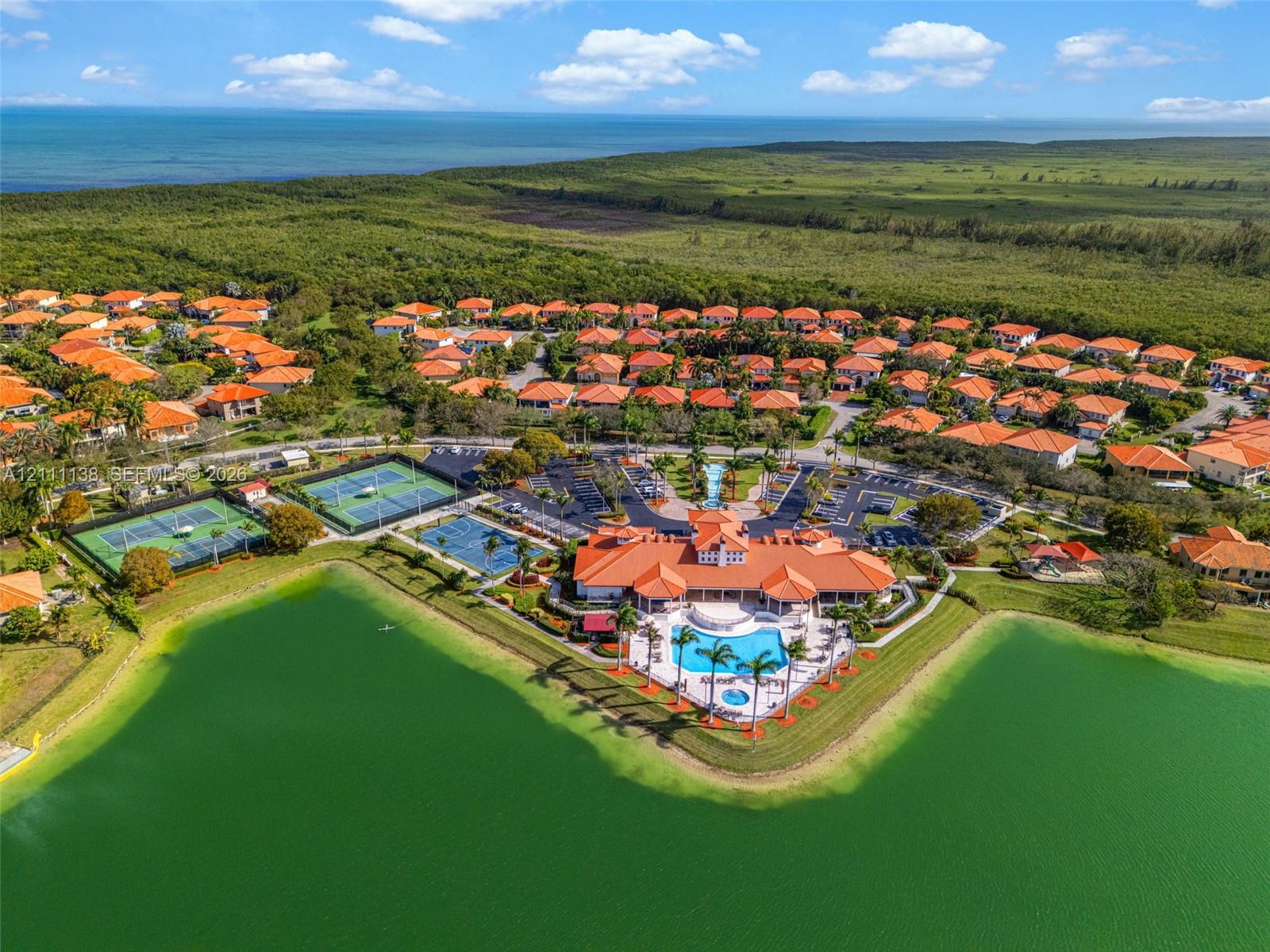 Aerial perspective of a community clubhouse featuring a swimming pool, multiple tennis courts, shoreline access, and proximity to the coast