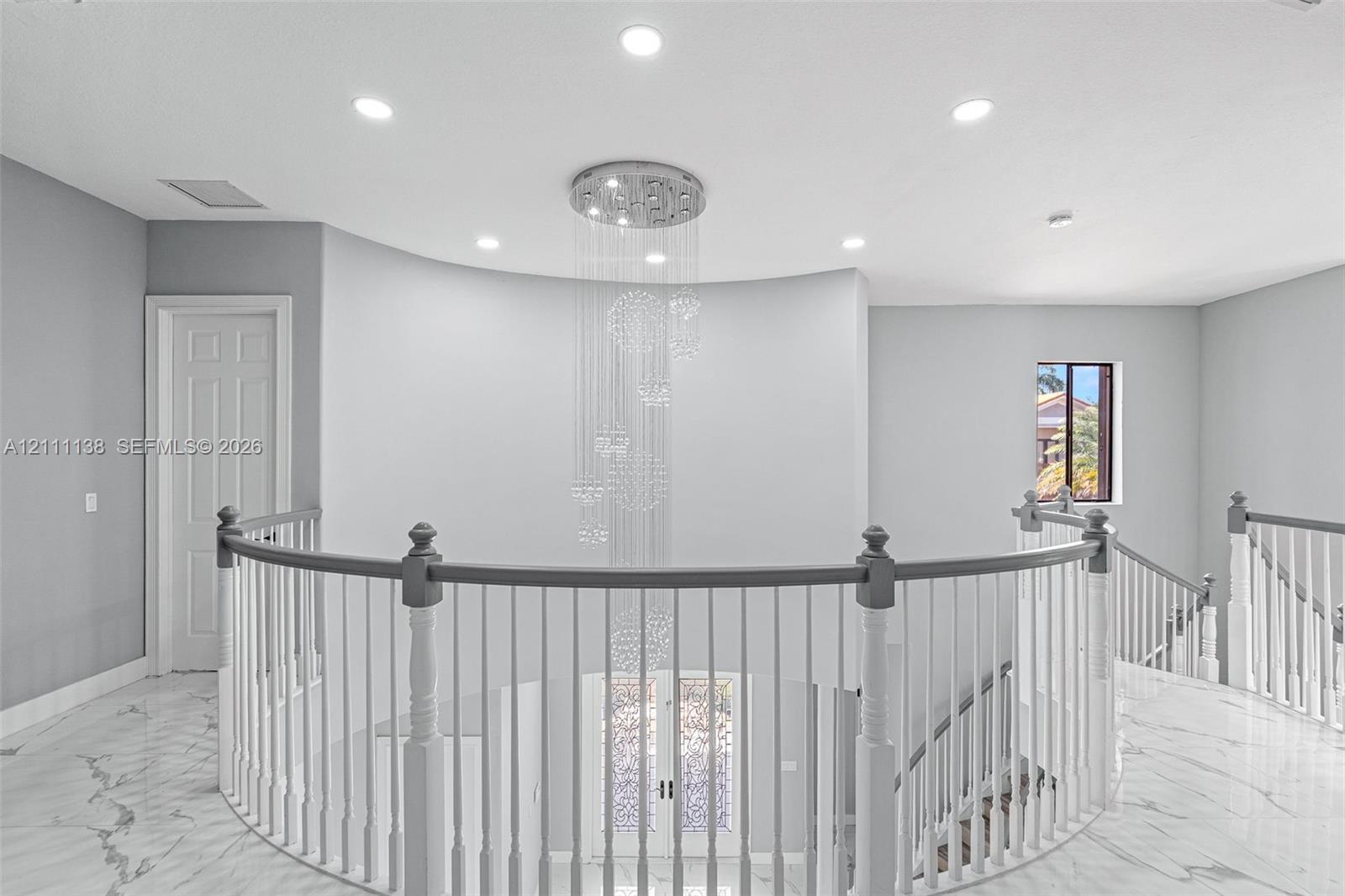 Curved architectural wall featuring a multi-tiered crystal chandelier, polished stone flooring, and a balustrade staircase with wood-finish handrails