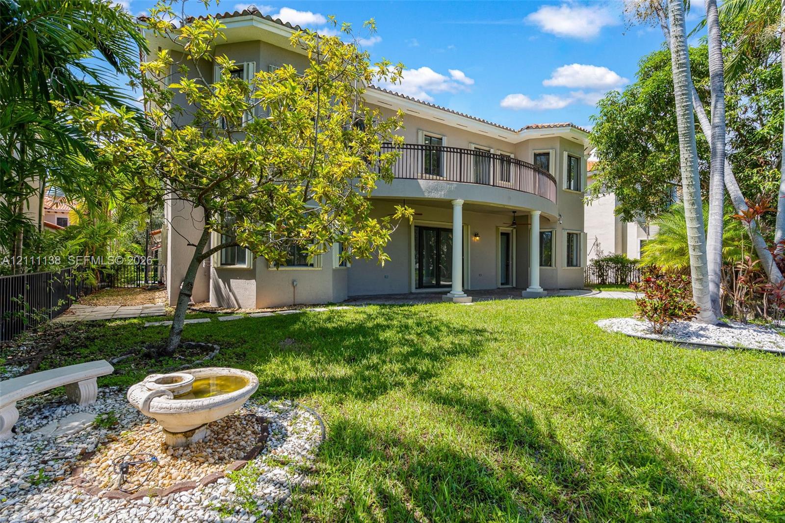 Two-story stucco exterior featuring a curved upper-level balcony with metal railing, classic columns, stone-accented landscaping, and a tiered fountain