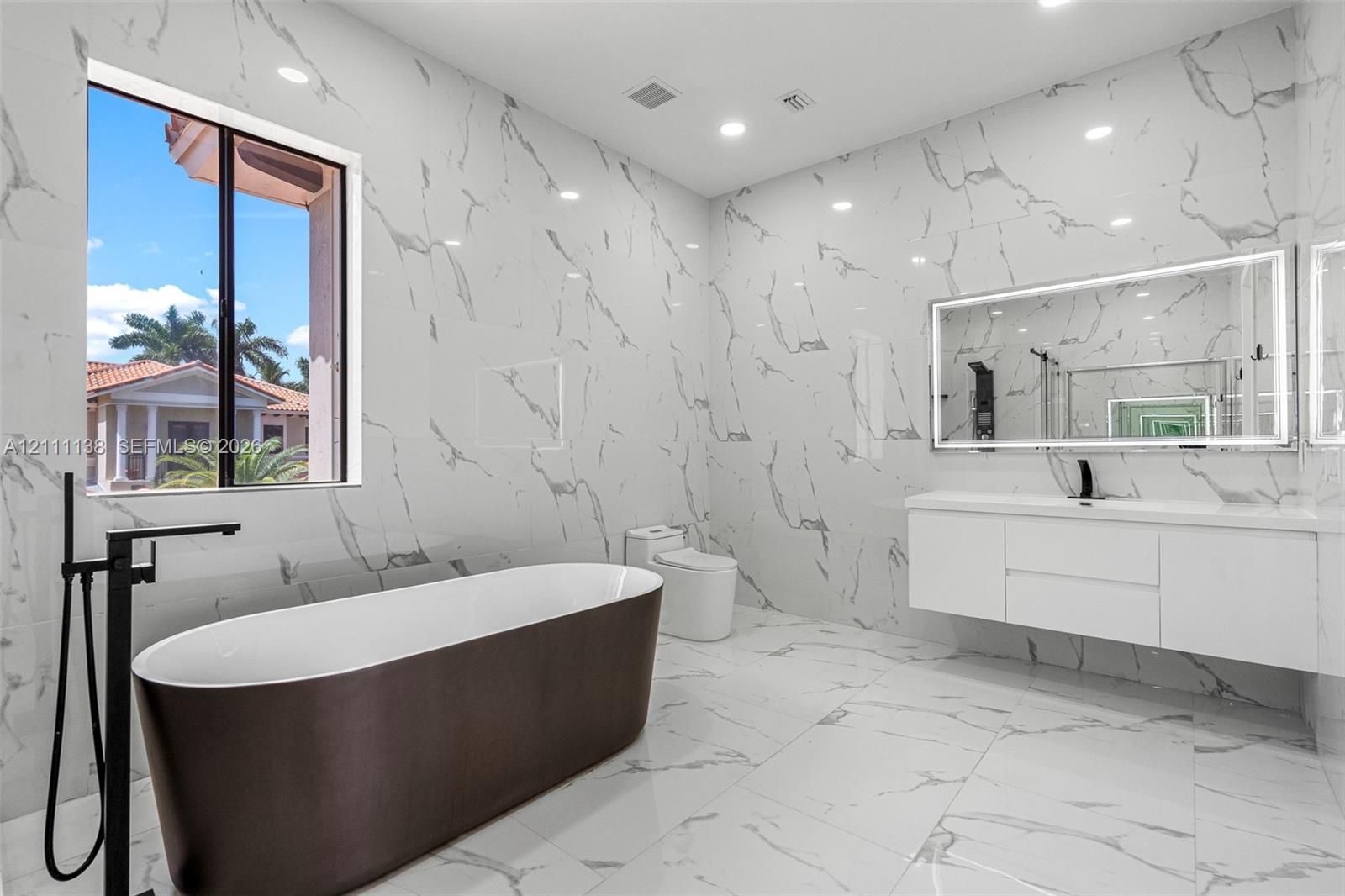 Bathroom featuring floor-to-ceiling large-format tile with stone-pattern finish, wall-mounted floating vanity, freestanding soaking tub with matte black floor-mounted faucet, and an oversized LED-backlit mirror