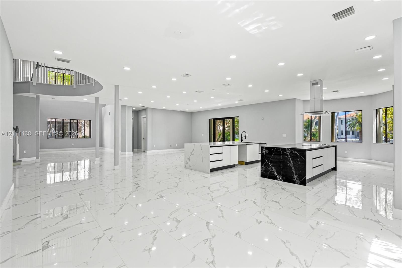 Open-concept living space featuring large-format tile flooring, dual kitchen islands with contrasting waterfall-edge stone surfaces, recessed ceiling lighting, and a curved staircase with a metal balustrade
