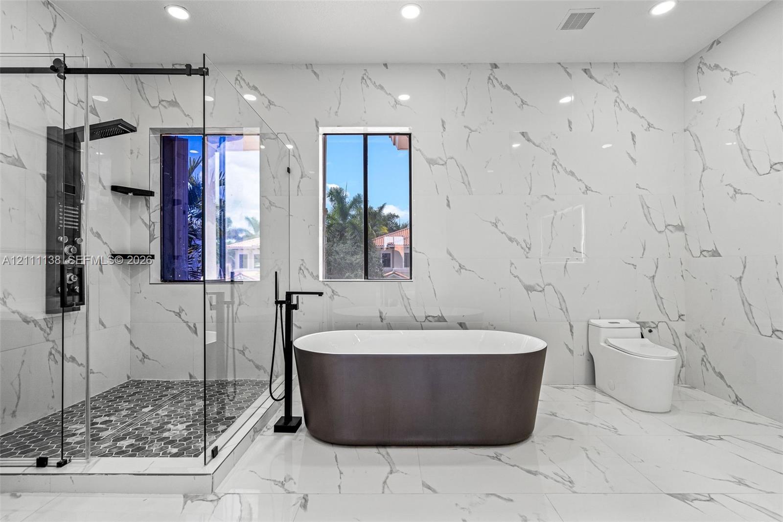 Full-height, large-format stone-patterned wall tile, freestanding soaking tub with floor-mounted matte black faucet, glass-enclosed walk-in shower with matte black rainfall showerhead, and hexagonal mosaic floor tile