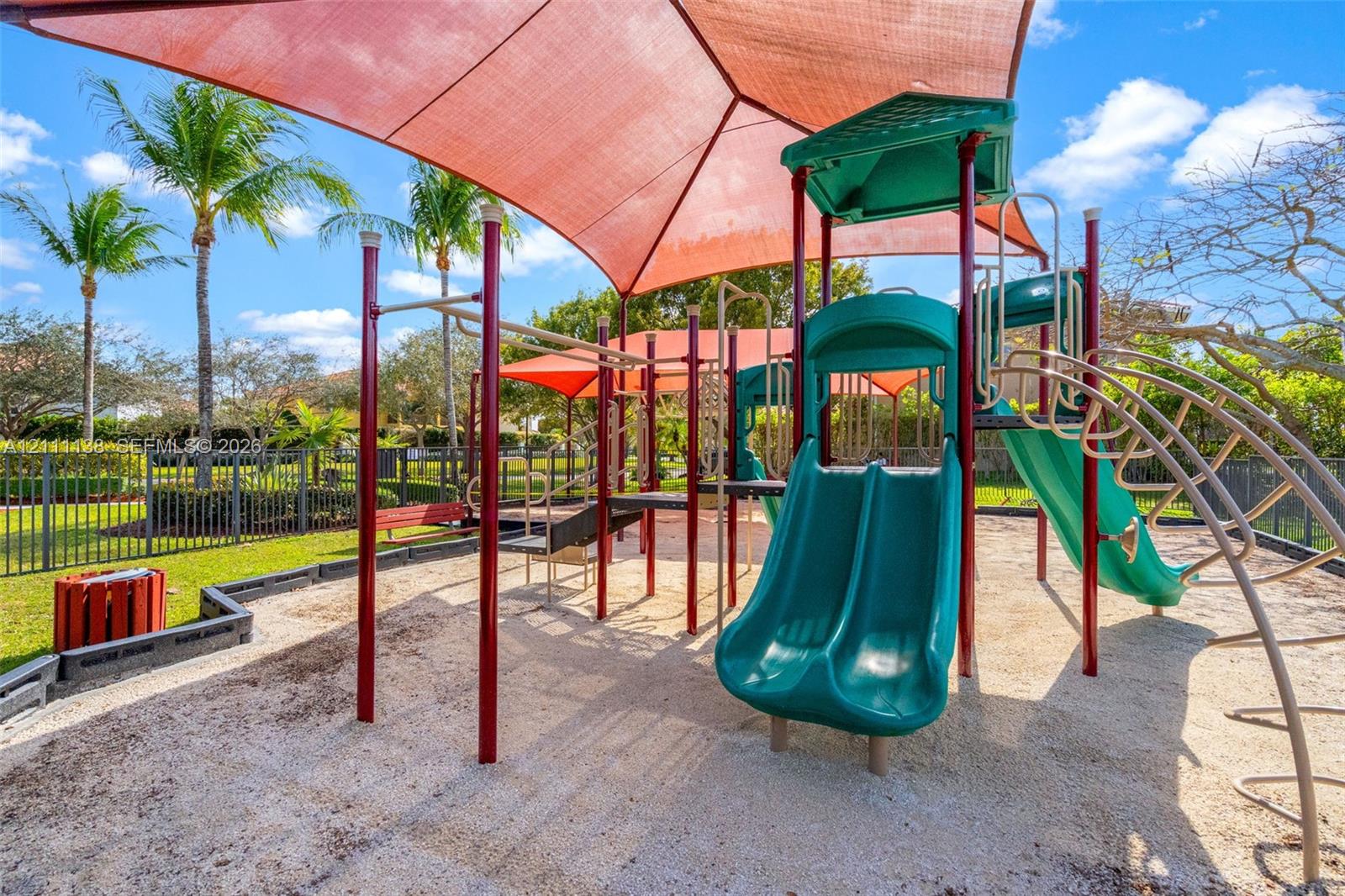 Outdoor recreational area featuring a multi-component play structure with dual slides, suspended shade canopy, rubberized mulch surfacing, and perimeter metal fencing