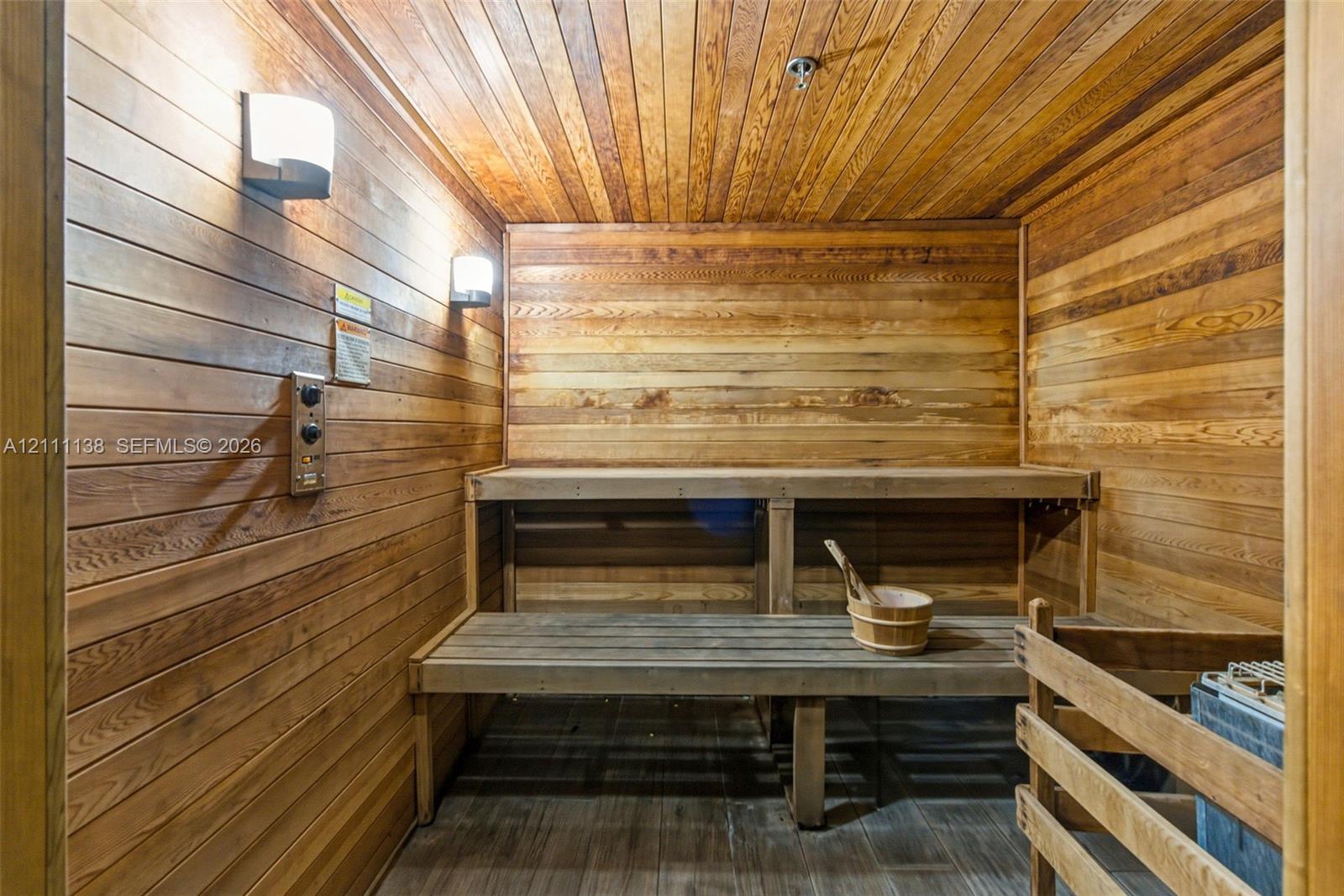 Sauna featuring wood paneling, multi-level built-in bench seating, recessed wall-mounted lighting, and a protective heater guard