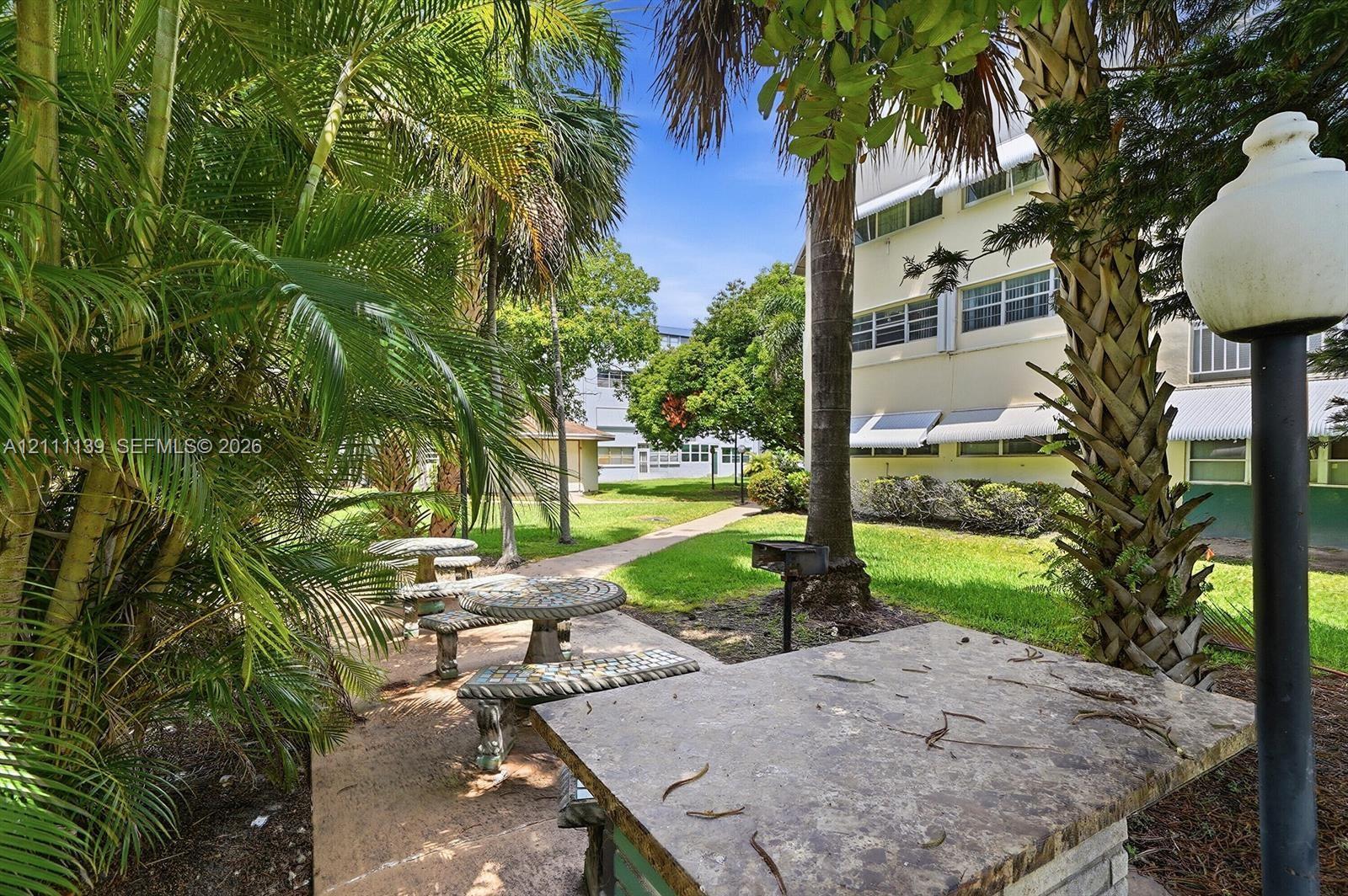 Common area featuring stone-finish picnic tables, a built-in outdoor grill, manicured lawn, and mature palm trees Common area featuring stone-finish picnic tables, a built-in outdoor grill, manicured lawn, and mature palm trees