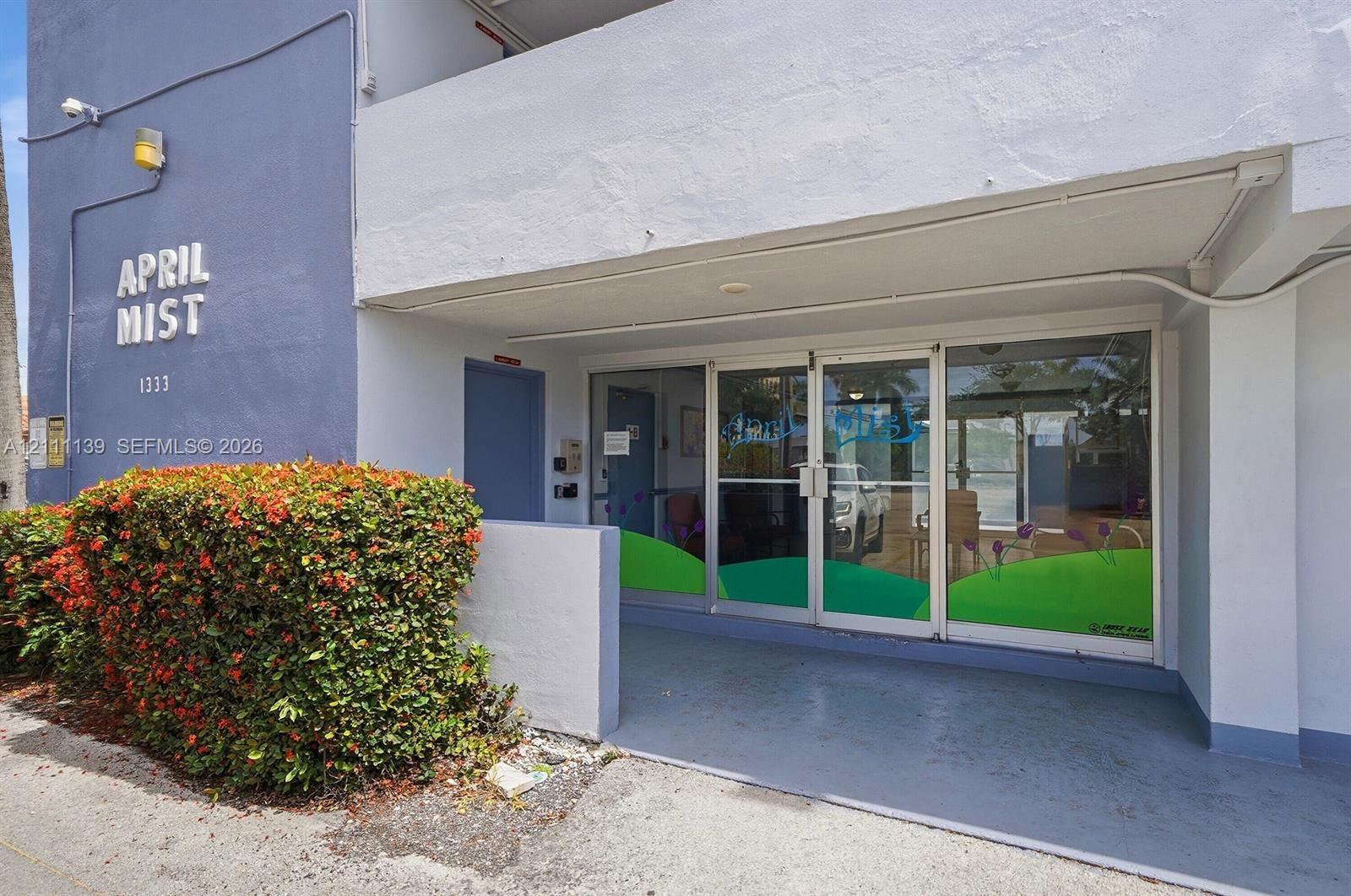 Exterior building entrance featuring a concrete facade, aluminum-framed glass storefront doors, a covered porch, and mature landscaping Exterior building entrance featuring a concrete facade, aluminum-framed glass storefront doors, a covered porch, and mature landscaping