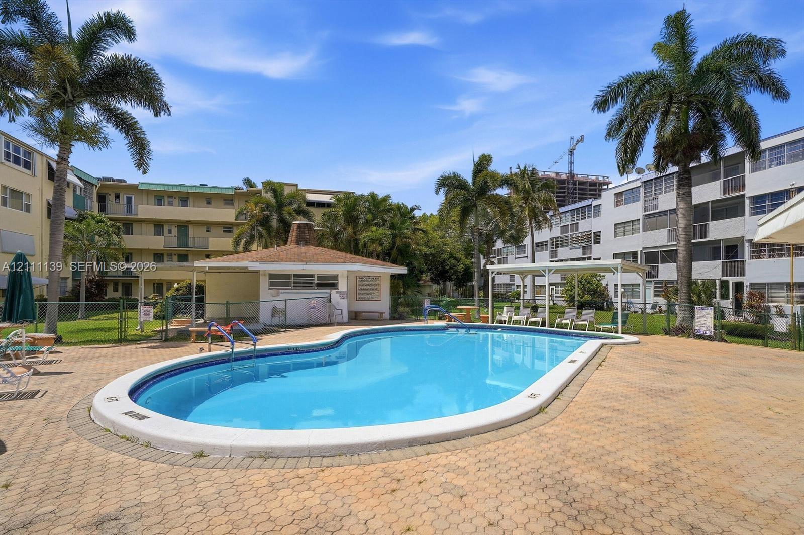 Community swimming pool featuring a concrete paver deck, surrounding palm trees, a covered patio structure, and a poolside cabana Community swimming pool featuring a concrete paver deck, surrounding palm trees, a covered patio structure, and a poolside cabana