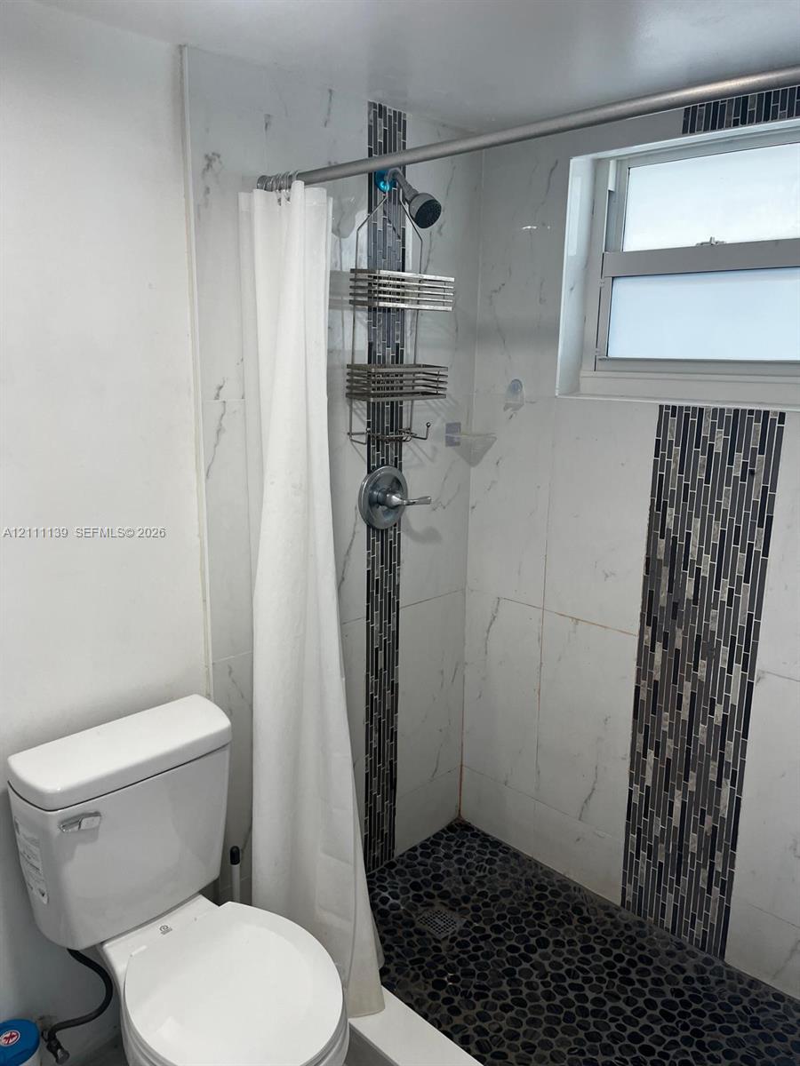 Shower featuring white wall tile with vertical mosaic accent inlay, pebble-style flooring, and a single-handle chrome fixture, paired with a standard-height porcelain toilet Shower featuring white wall tile with vertical mosaic accent inlay, pebble-style flooring, and a single-handle chrome fixture, paired with a standard-height porcelain toilet