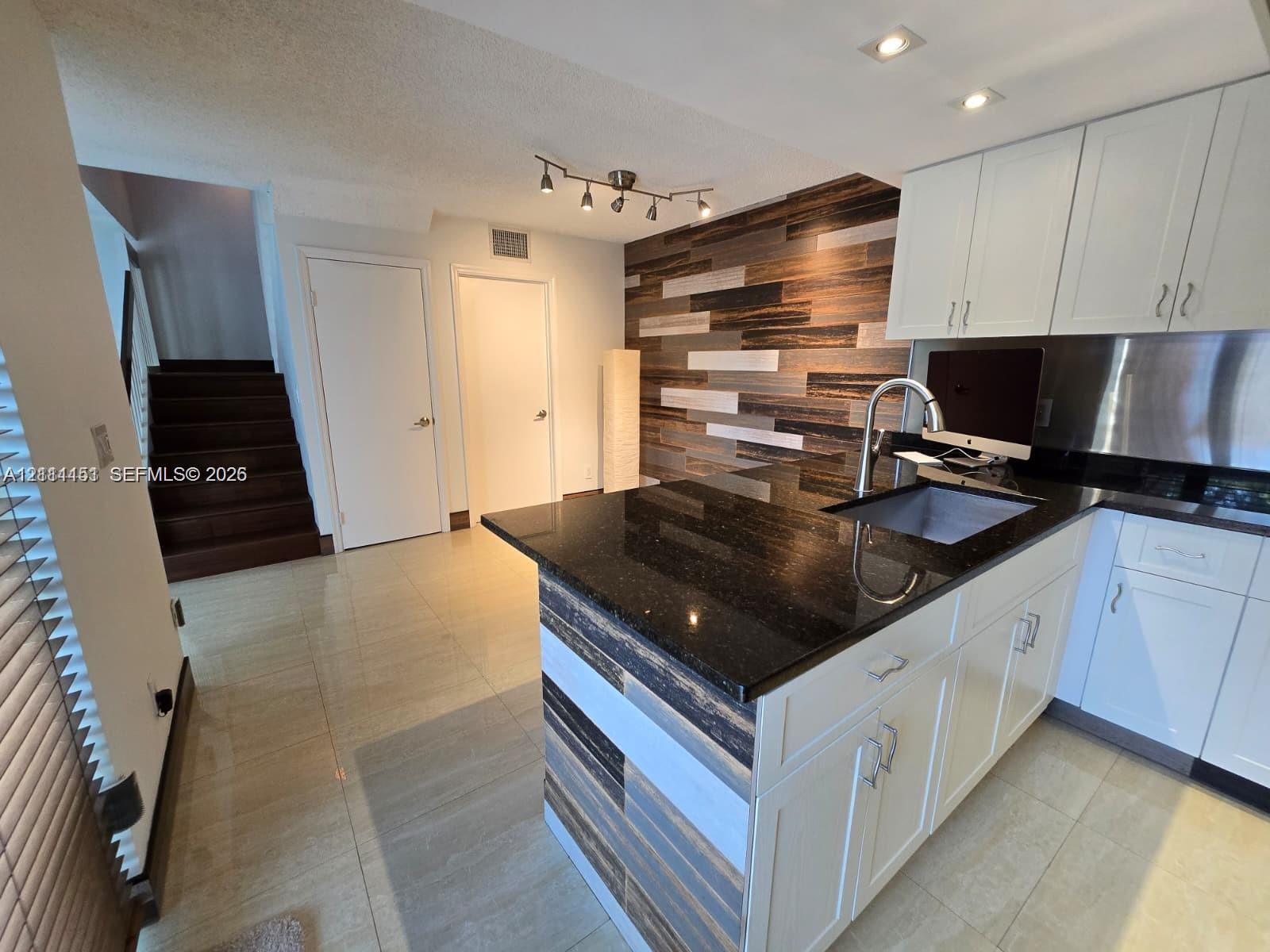 Kitchen island featuring a dark stone countertop, undermount sink, and multi-toned wood-finish paneling, complemented by white cabinetry, stainless steel backsplash, and a decorative wood-finish wall accent Kitchen island featuring a dark stone countertop, undermount sink, and multi-toned wood-finish paneling, complemented by white cabinetry, stainless steel backsplash, and a decorative wood-finish wall accent