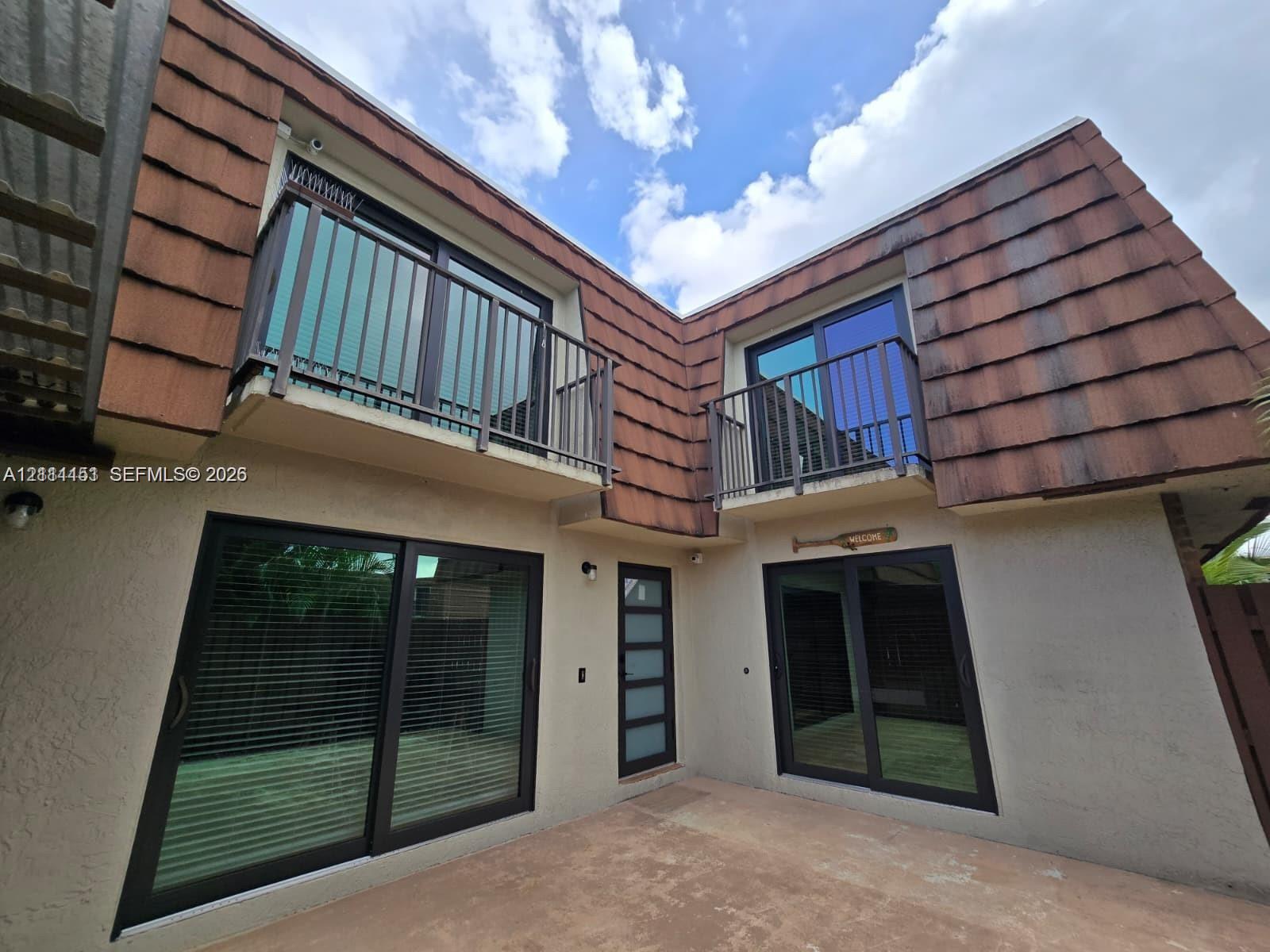 Exterior featuring dual upper-level balconies with metal railings, sliding glass doors with dark frames, a modern entry door with frosted glass panels, and textured shingled roof accents Exterior featuring dual upper-level balconies with metal railings, sliding glass doors with dark frames, a modern entry door with frosted glass panels, and textured shingled roof accents