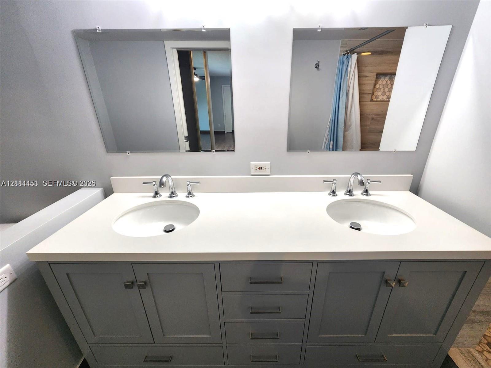 Double vanity featuring a light-toned solid surface countertop, dual undermount sinks, chrome widespread faucets, and gray Shaker-style cabinetry with matte finish hardware, topped by two frameless wall-mounted mirrors