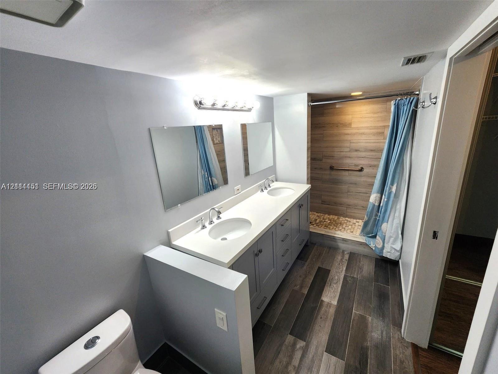 Dual-sink vanity with a solid surface countertop, rectangular mirrors, wood-finish floor tiling, and a walk-in shower featuring horizontal tile wall accents and stone-style flooring