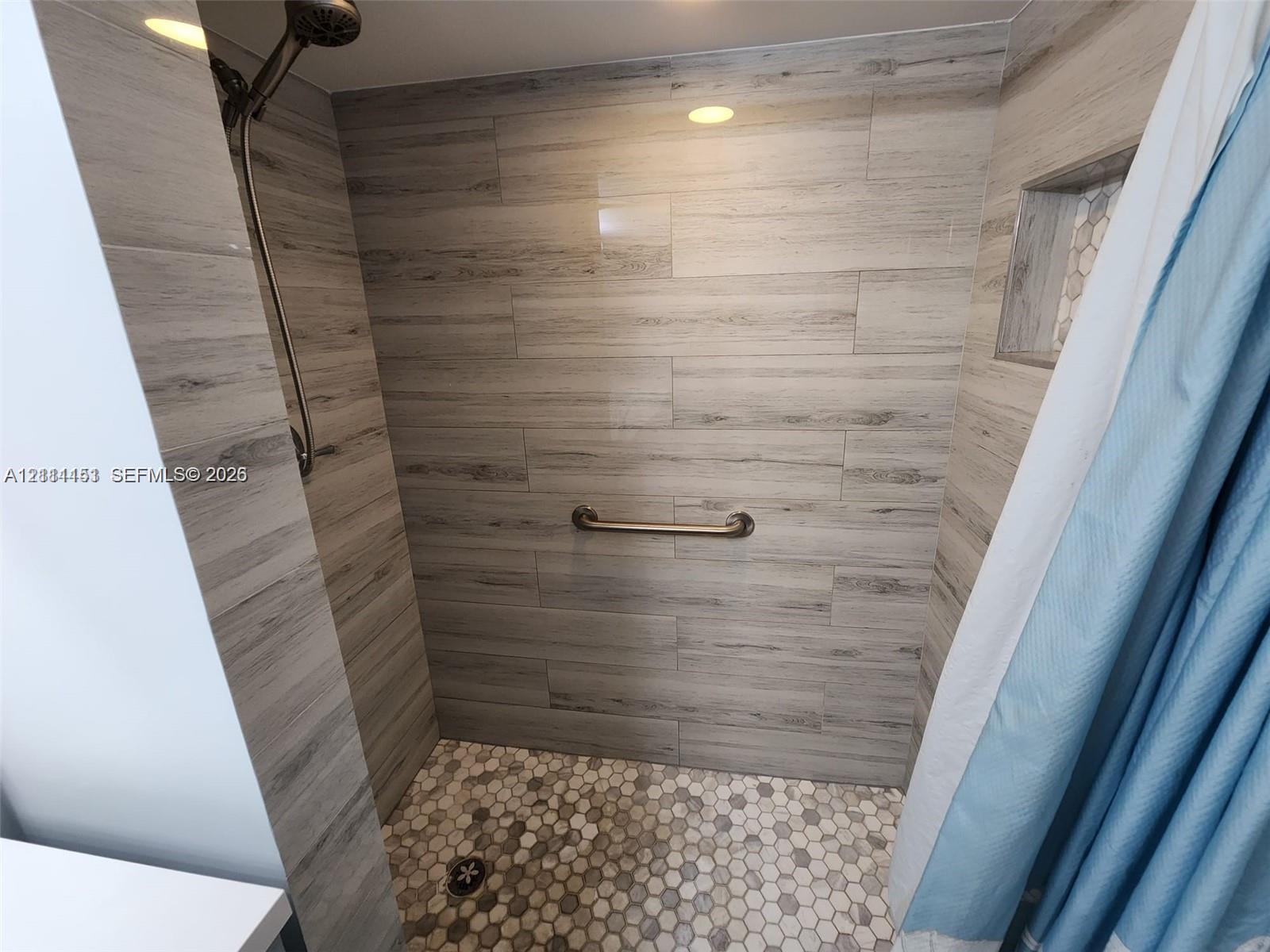 Walk-in shower with wood-finish tile wall surround, hexagonal floor tiling, recessed wall niche, stainless steel shower head, and wall-mounted grab bar