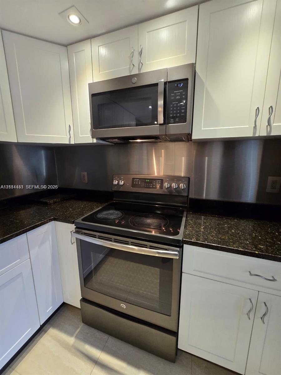 Kitchen featuring white shaker-style cabinetry, dark stone countertops, a full-height stainless steel backsplash, and matching stainless steel electric range and microwave