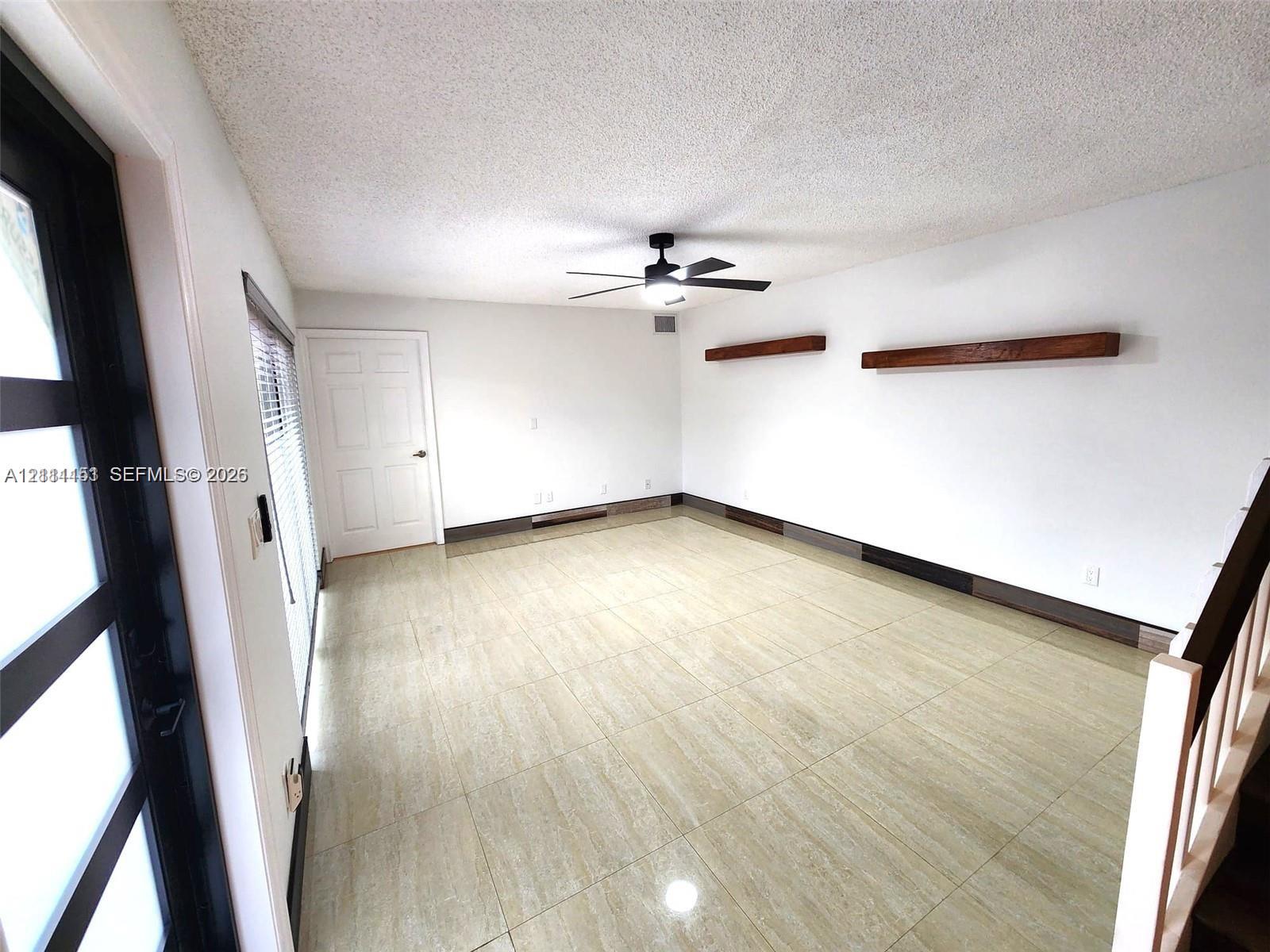 Neutral-toned tile flooring, dual wooden floating shelves, black-framed glass entry door, and a central ceiling fan with integrated light fixture