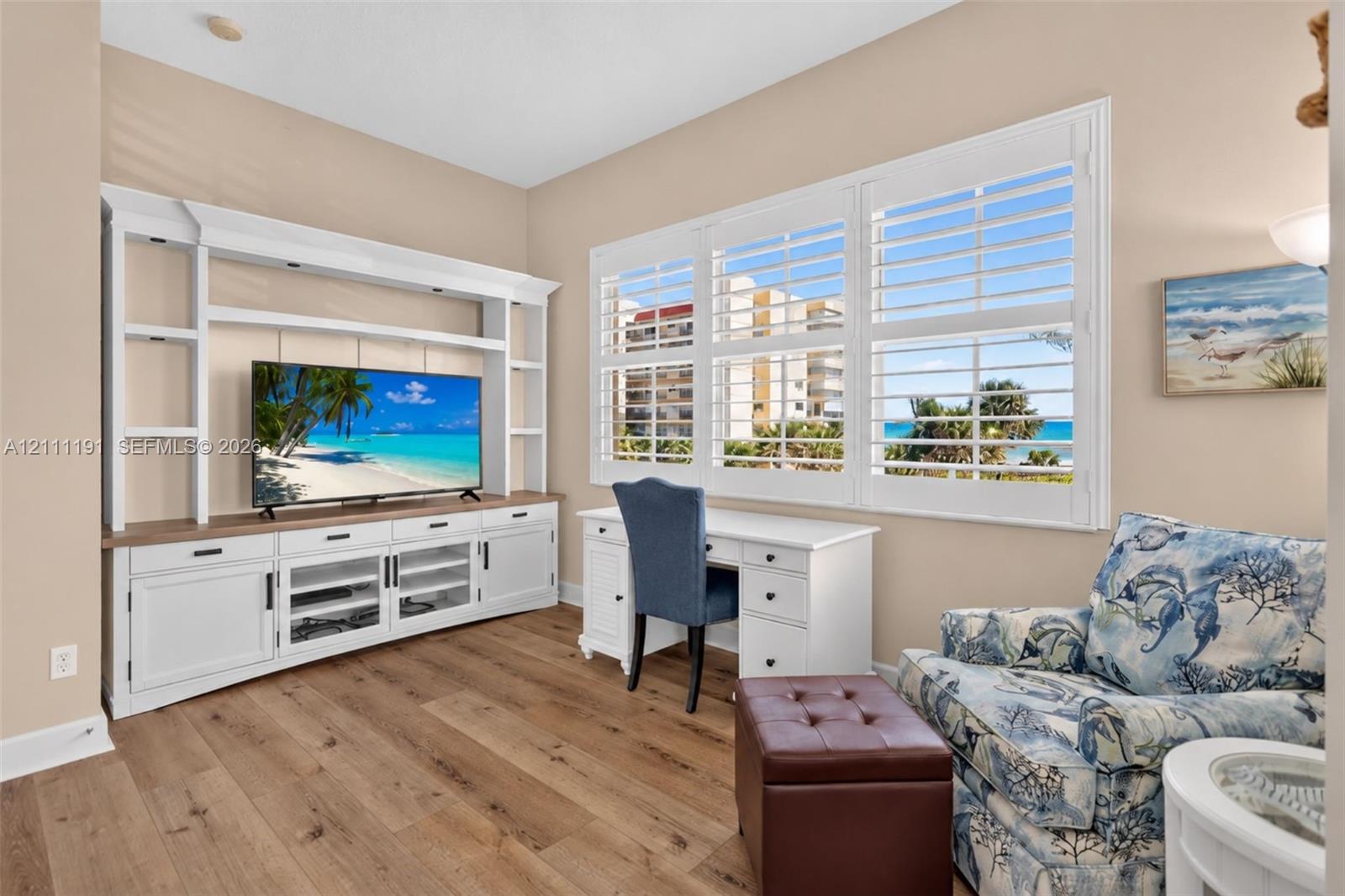 Built-in media console with white cabinetry, wood-finish flooring, and white plantation shutters framing coastal scenery