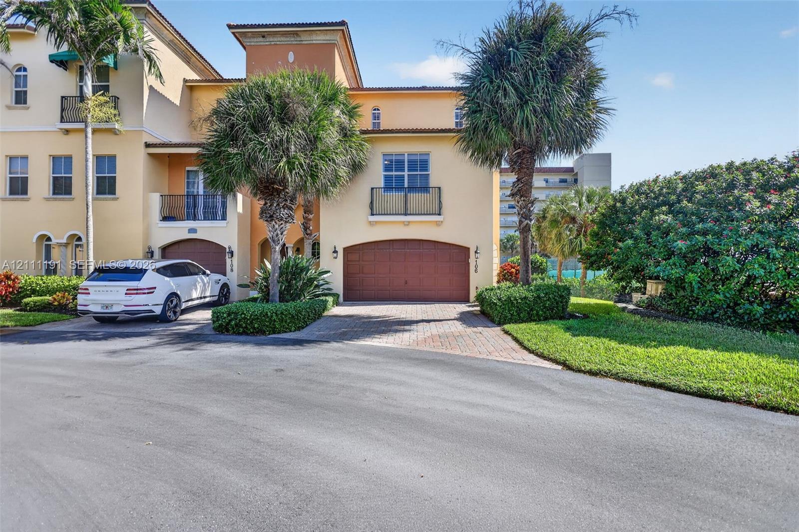 Mediterranean-inspired exterior featuring stucco facade, arched garage doors, upper-level balconies with metal railings, paver driveway, and mature landscaping