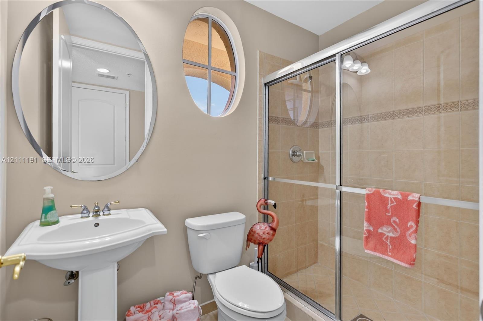 Entry-level Bathroom featuring a pedestal sink, oval wall mirror, shower with sliding glass doors and tiled surround, and a fixed arched window