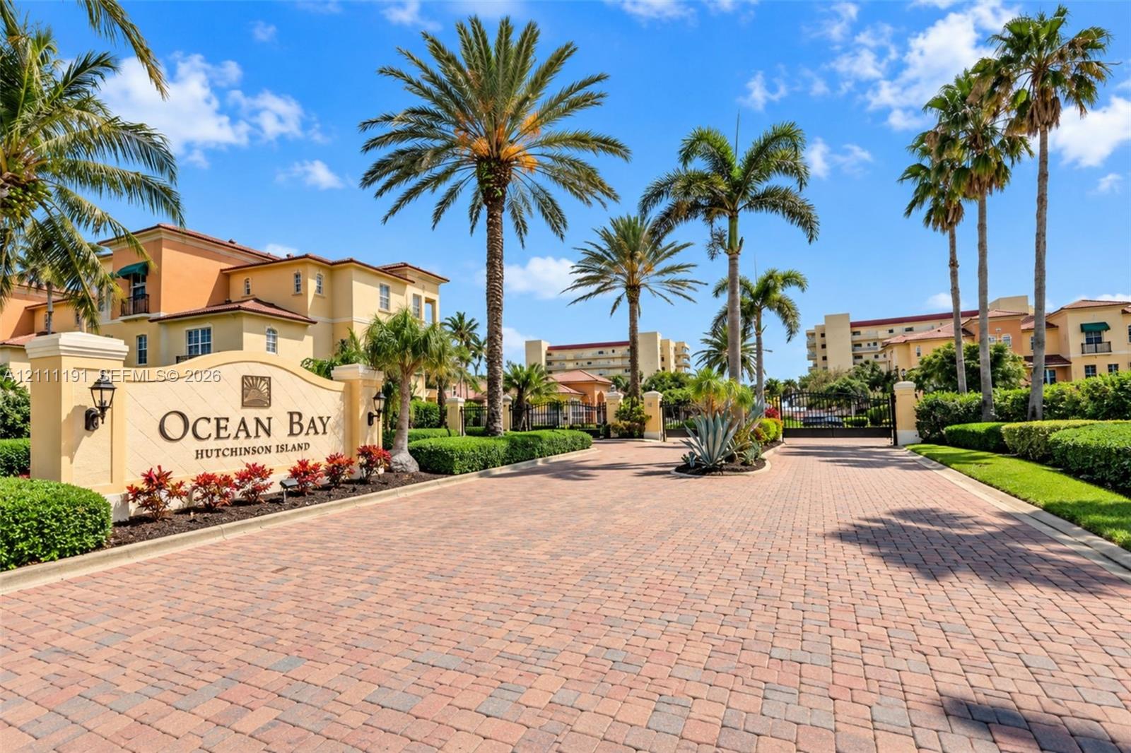 Gated entrance featuring a brick-paver drive, custom stone monument signage, mature palm landscaping, and integrated wall-mounted lighting Gated entrance featuring a brick-paver drive, custom stone monument signage, mature palm landscaping, and integrated wall-mounted lighting