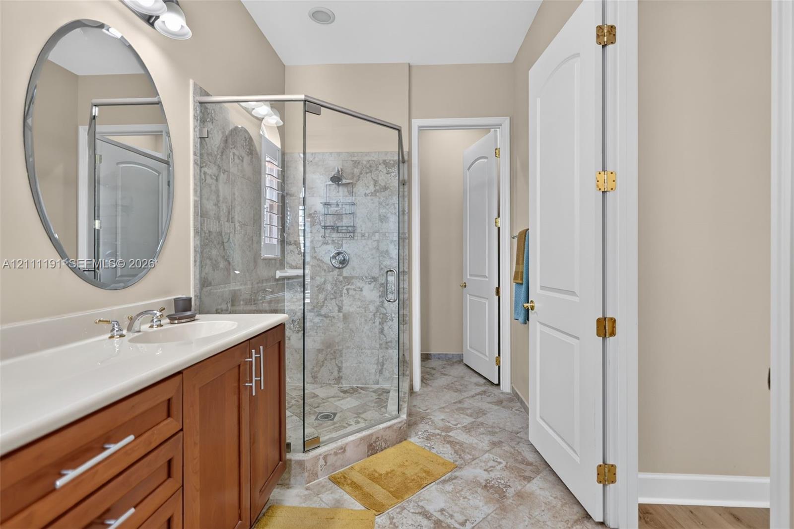 Primary Bathroom featuring a wood-finish vanity with a white countertop and oval mirror, walk-in shower with glass enclosure and tiled walls, and ceramic tile flooring. Water closet is behind shower.