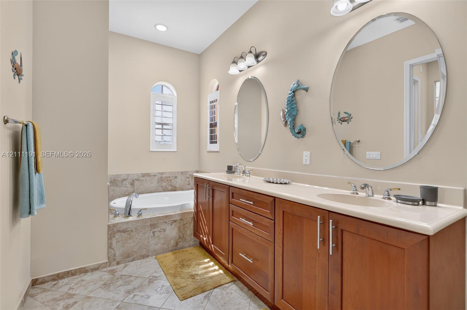 Primary Bathroom. Dual-sink vanity with wood-finish cabinetry, solid surface countertop, inset soaking tub, tile surround, and oval wall-mounted mirrors