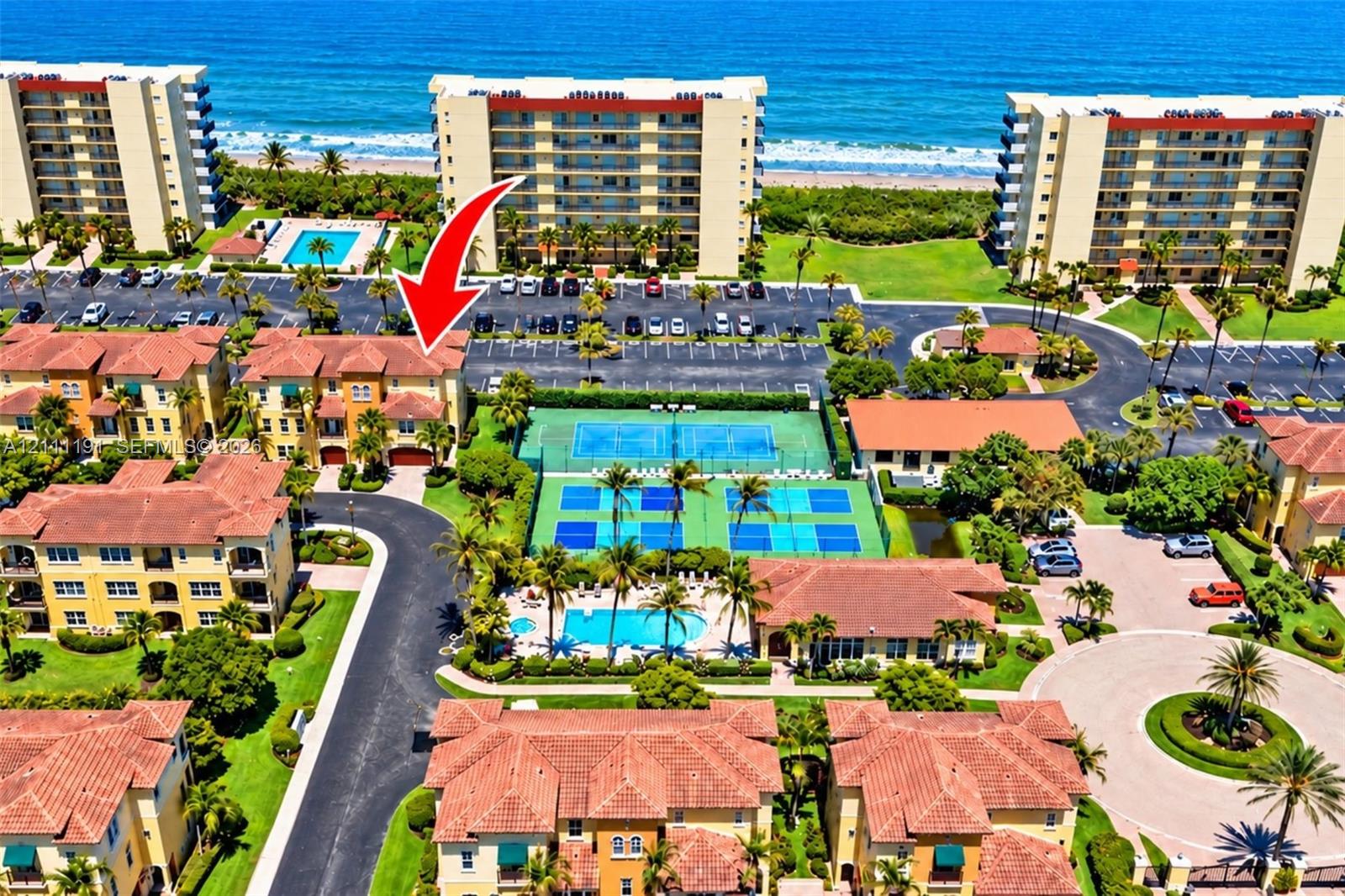 Aerial perspective of a multi-story residential building featuring a tile roof, dedicated parking, tennis courts, and a community pool