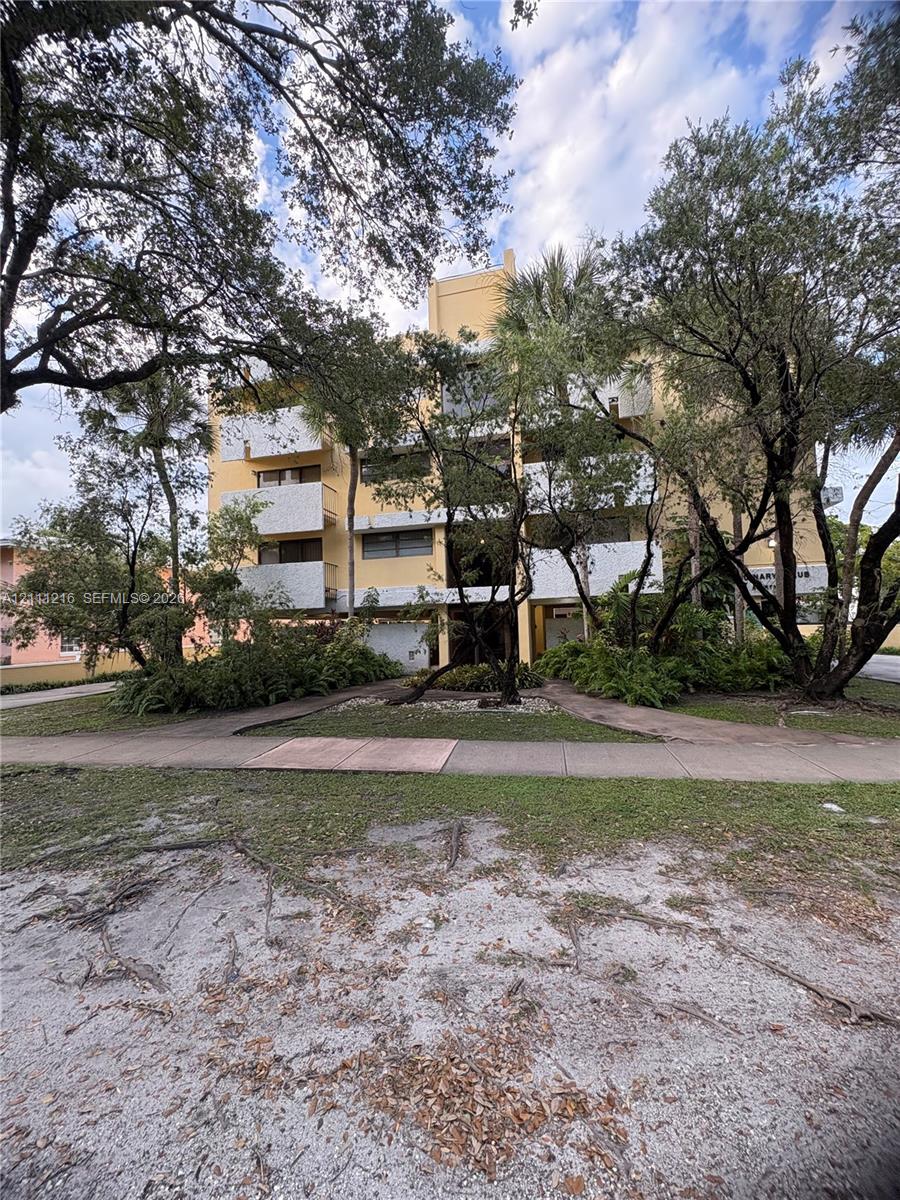 Multi-story building exterior featuring concrete construction, white balcony railings, and mature canopy trees