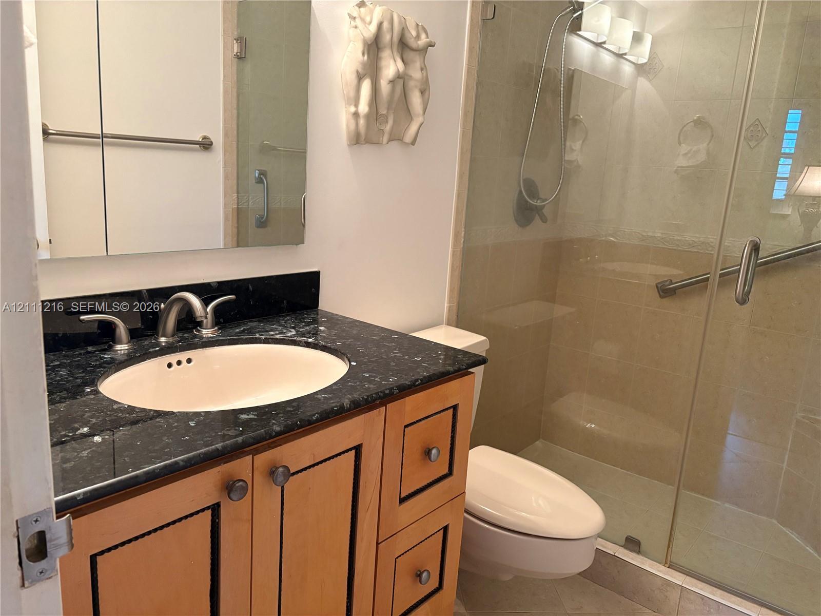 Wood-finish vanity with a stone countertop, oval undermount sink, glass-enclosed shower, and wall-mounted vanity lighting