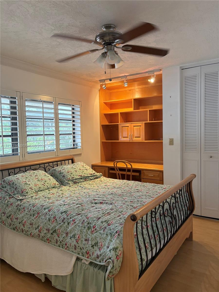Built-in wood shelving with integrated desk, white louvered-style closet doors, wood-finish flooring, plantation shutters, and ceiling fan with integrated lighting