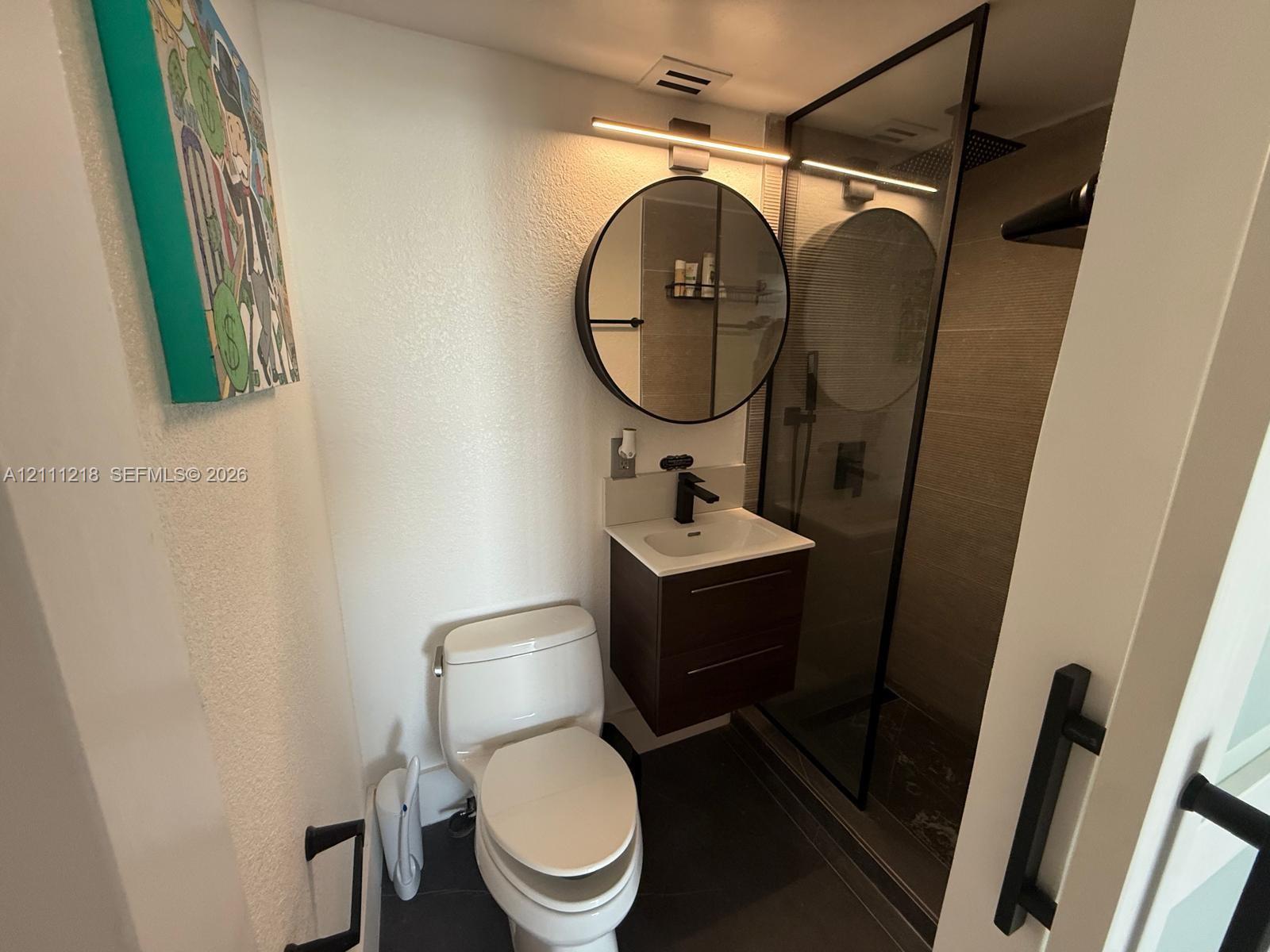 Modern floating vanity with a light-toned countertop and matte black faucet, paired with a circular mirror, linear vanity lighting, and a glass-enclosed shower with tiled surround