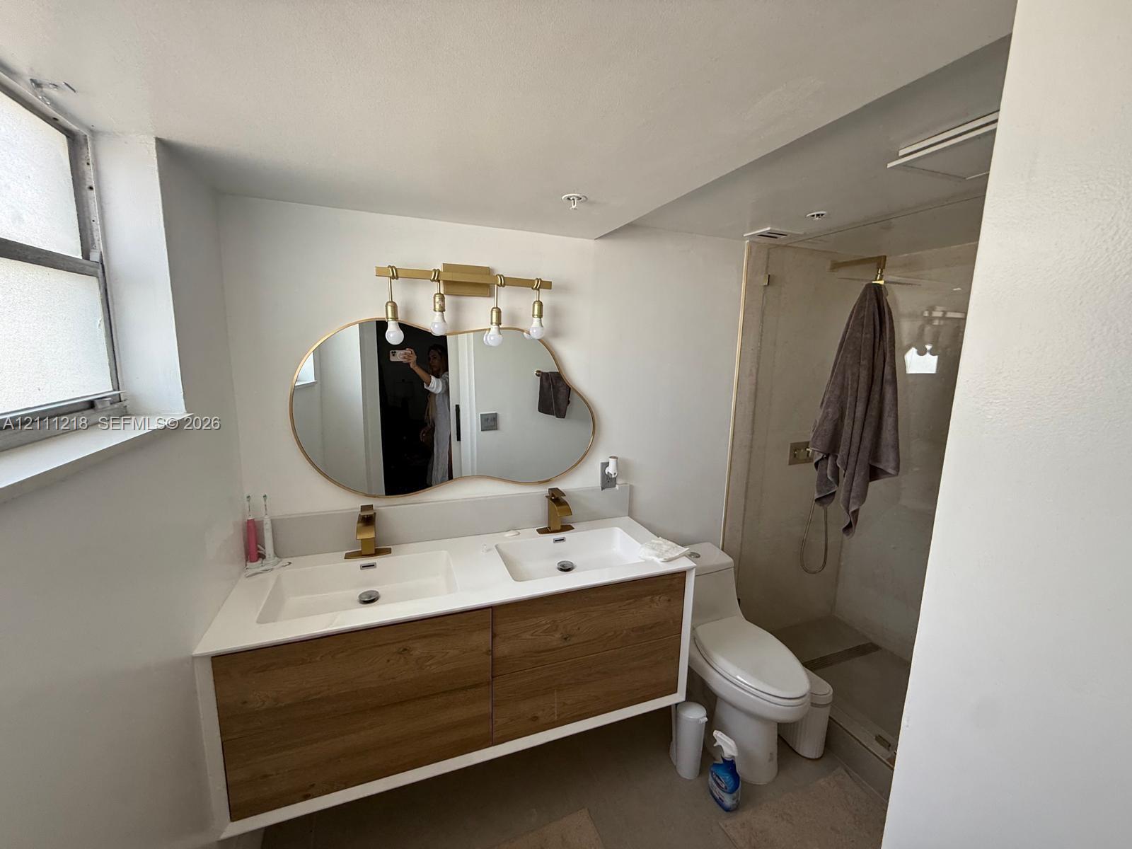 Floating dual-sink vanity with wood-finish cabinetry, quartz countertop, brass-toned fixtures, organic-shaped mirror, and glass-enclosed shower