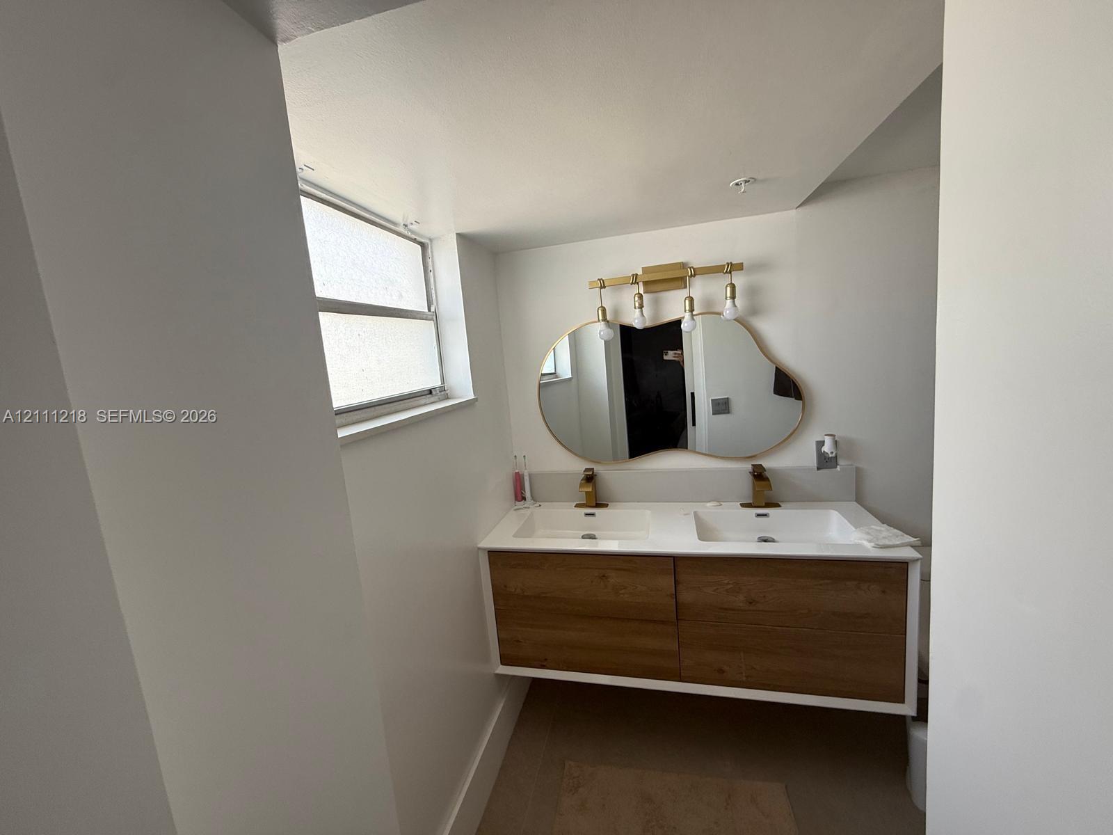 Floating wood-finish vanity with dual integrated sinks, metallic hardware, an organic-shaped wall mirror, and modern multi-bulb lighting