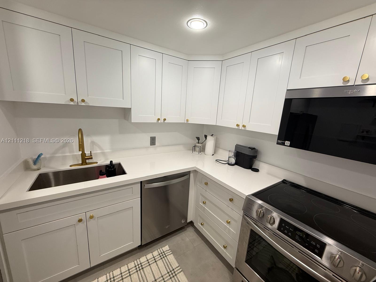 Kitchen featuring white shaker-style cabinetry, solid-surface countertops, stainless steel appliances, and brass-tone hardware Kitchen featuring white shaker-style cabinetry, solid-surface countertops, stainless steel appliances, and brass-tone hardware