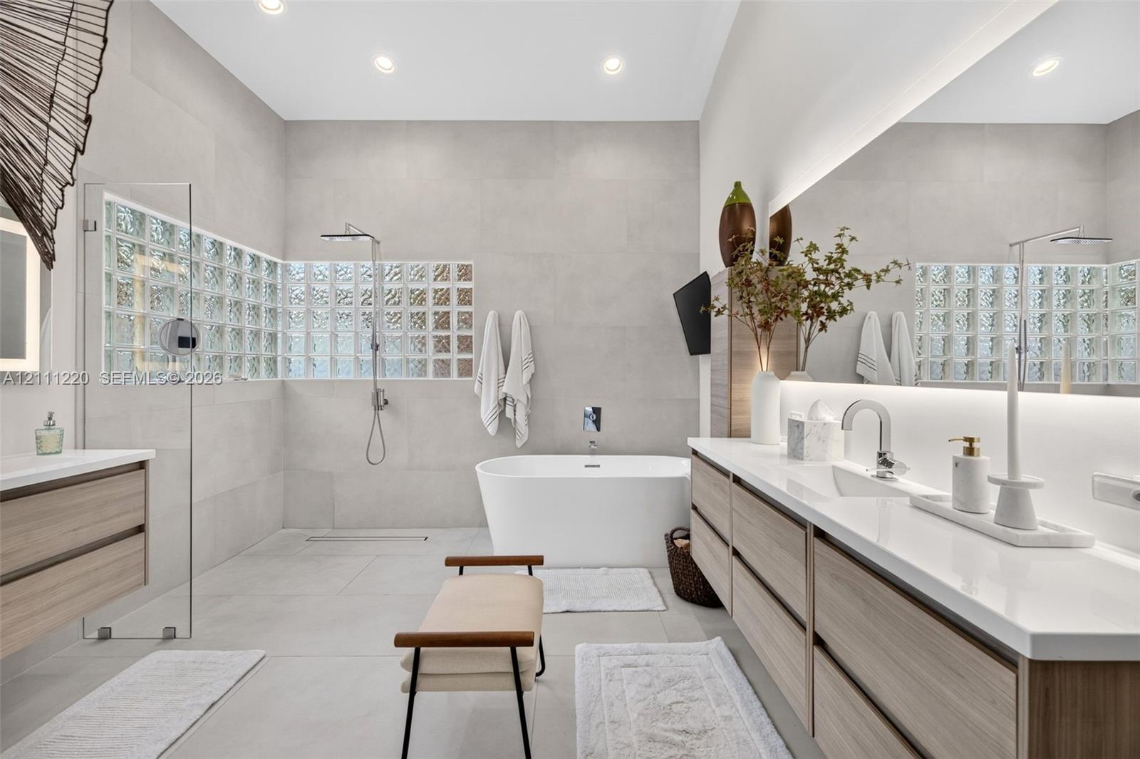 Bathroom featuring a large glass-enclosed shower, freestanding soaking tub, dual-sink vanity with light wood-finish cabinetry, quartz countertops, and back-lit wall mirror