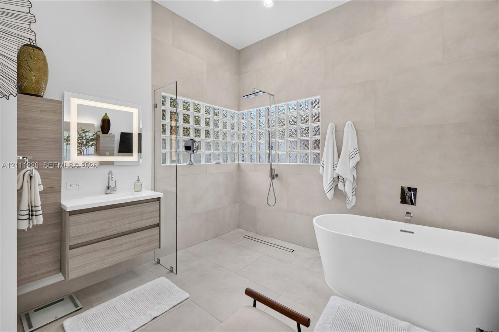 Bathroom featuring a freestanding soaking tub, walk-in shower with glass partition, neutral large-format wall tile, wood-finish wall-mounted vanity, and glass block accent windows