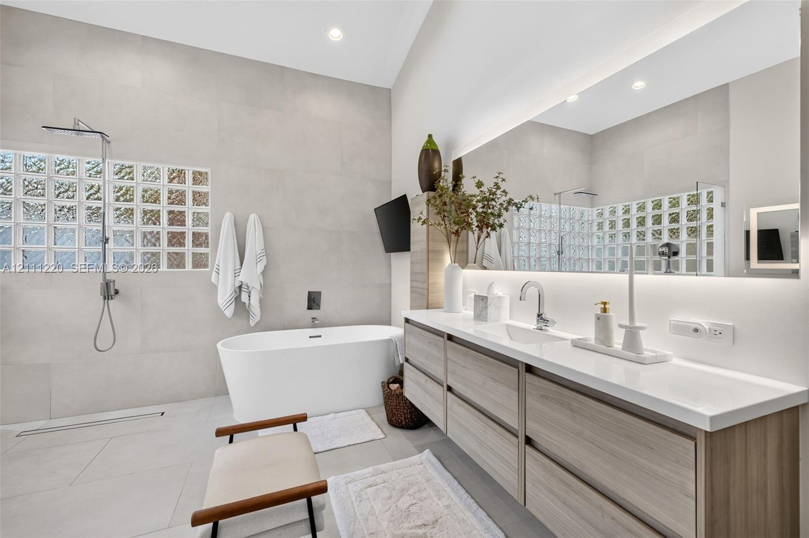 Bathroom featuring a floating wood-finish vanity with a countertop basin, a standalone soaking tub, full-wall tiling, a glass-block privacy window, and an expansive backlit mirror