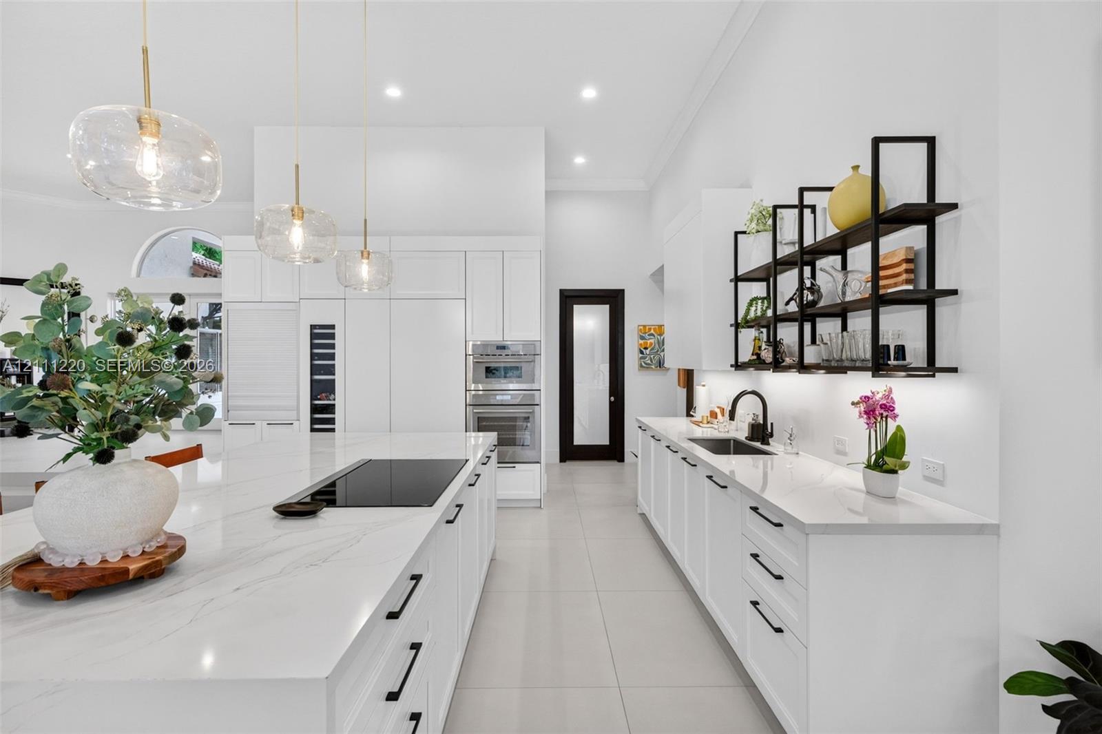 Modern kitchen featuring a stone-surface island with an integrated induction cooktop, white cabinetry with matte black hardware, stainless steel double wall ovens, glass pendant lighting, and black metal open shelving
