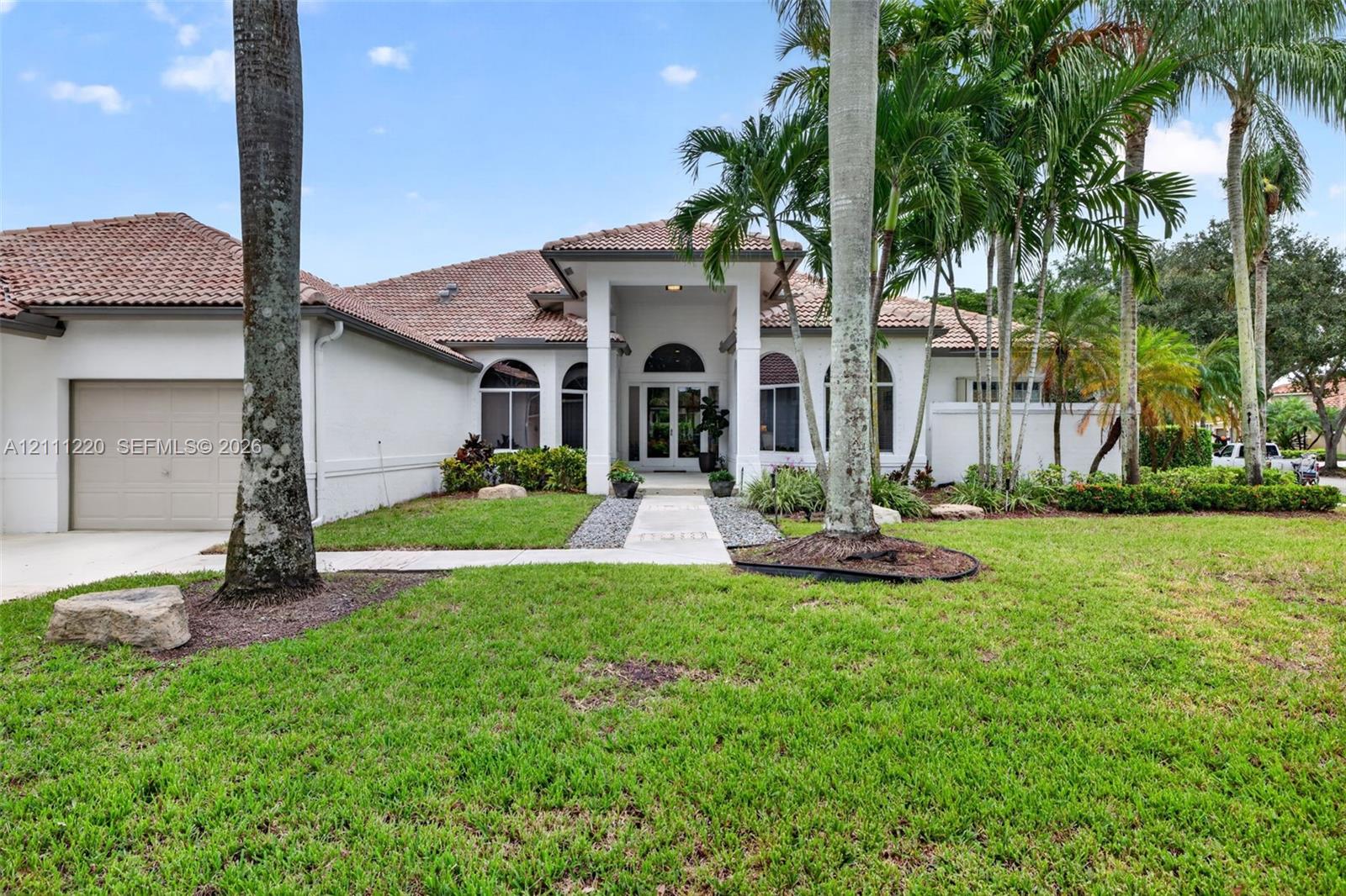 Stucco exterior featuring a tile roof, arched windows, covered entryway, and manicured landscaping with mature palm trees Stucco exterior featuring a tile roof, arched windows, covered entryway, and manicured landscaping with mature palm trees