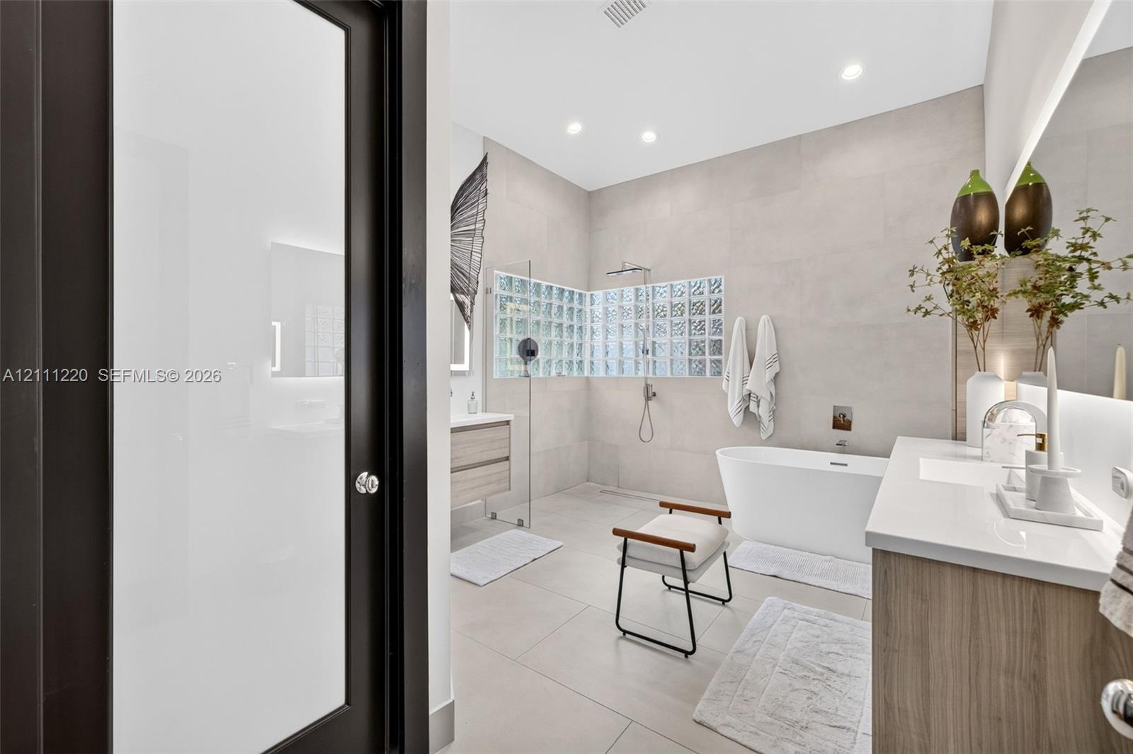 Bathroom featuring large-format tile flooring and walls, a freestanding soaking tub, a wet-room style shower enclosure with glass block wall detailing, and a wood-finish vanity with an integrated countertop