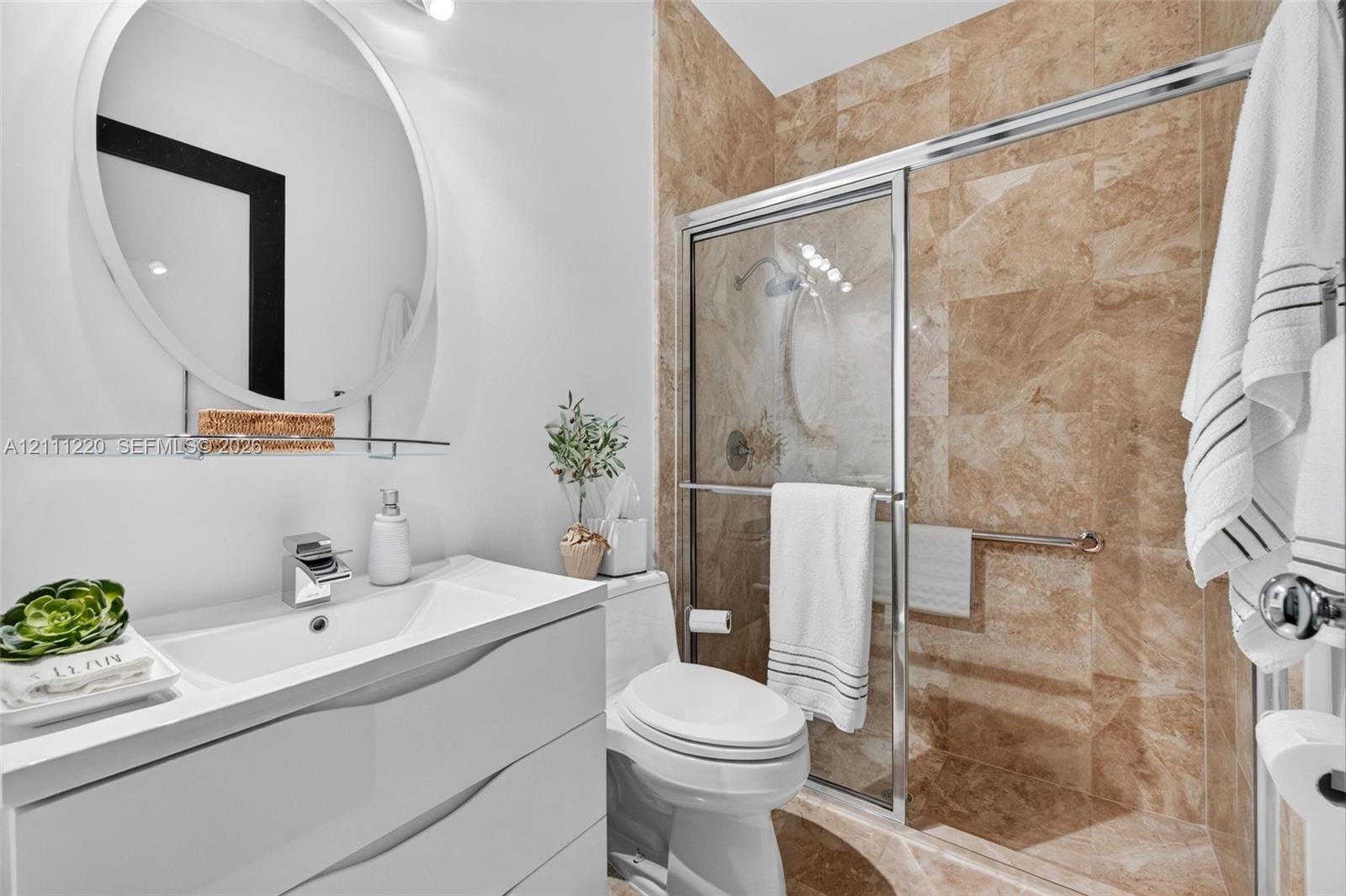 Contemporary white vanity with integrated basin, polished chrome faucet, circular wall mirror, and walk-in shower featuring floor-to-ceiling stone-look tiling and sliding glass enclosure