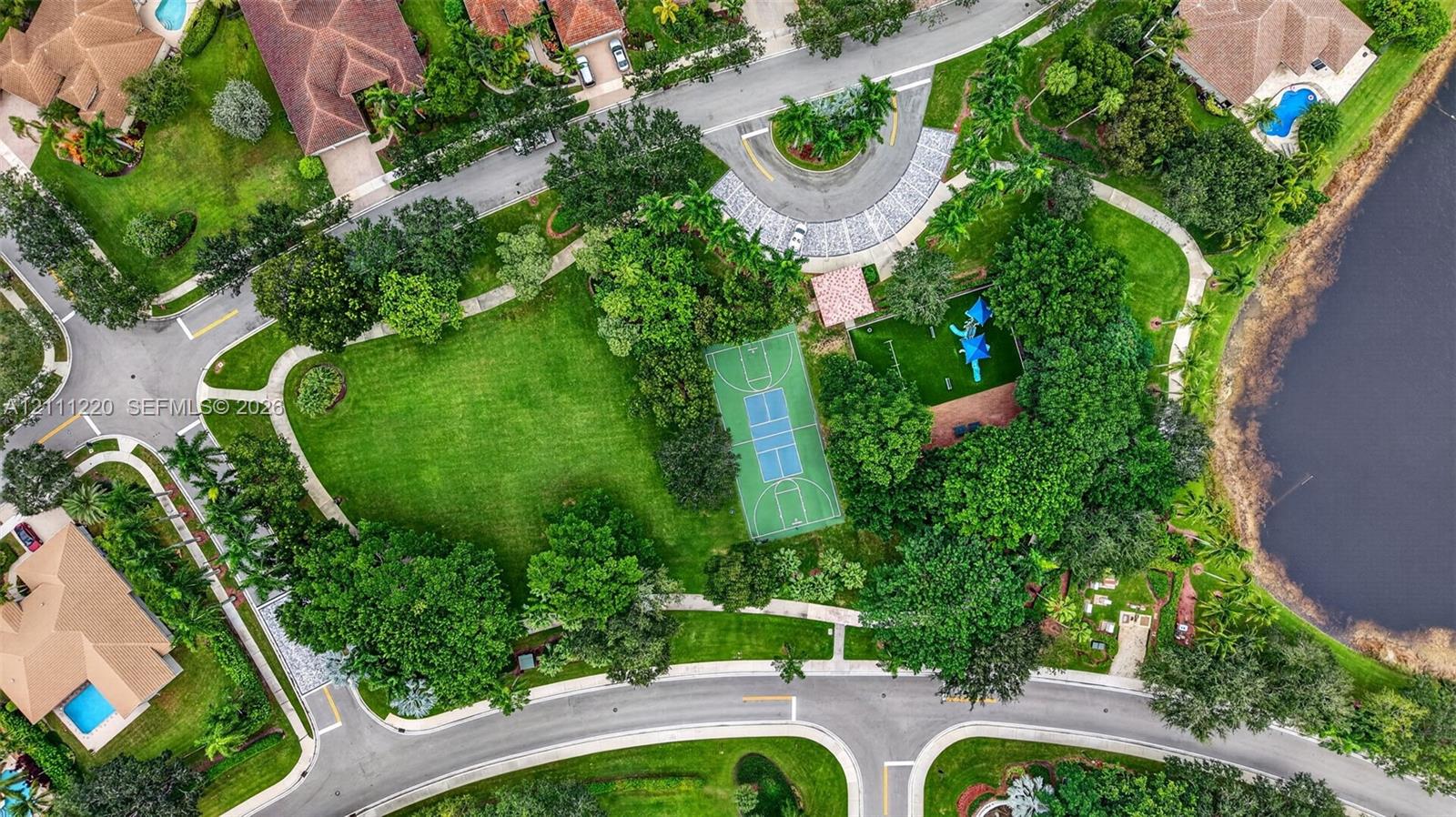 Aerial perspective of a community recreation area featuring a basketball court, playground, expansive green space, mature trees, and a bordering water feature