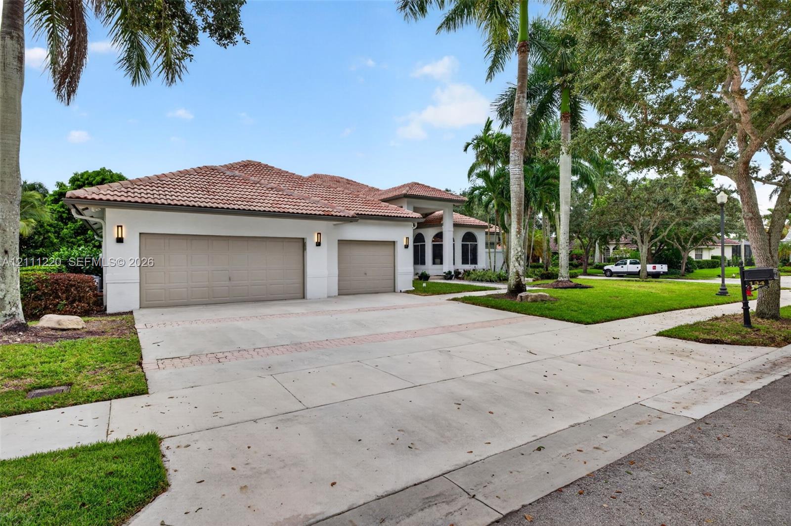 Stucco exterior featuring a multi-gable tile roof, three-car garage, concrete driveway with brick inlay, and mature landscaping