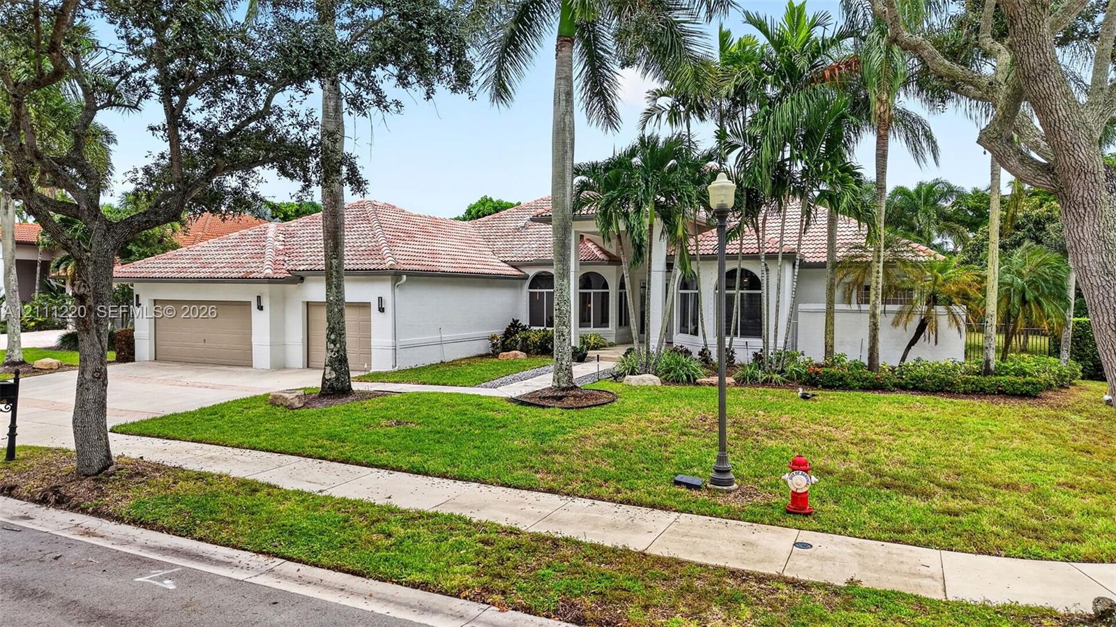 Single-story stucco exterior featuring a multi-car garage, arched windows, and a tiled roof, set within a landscaped lot with mature palm trees